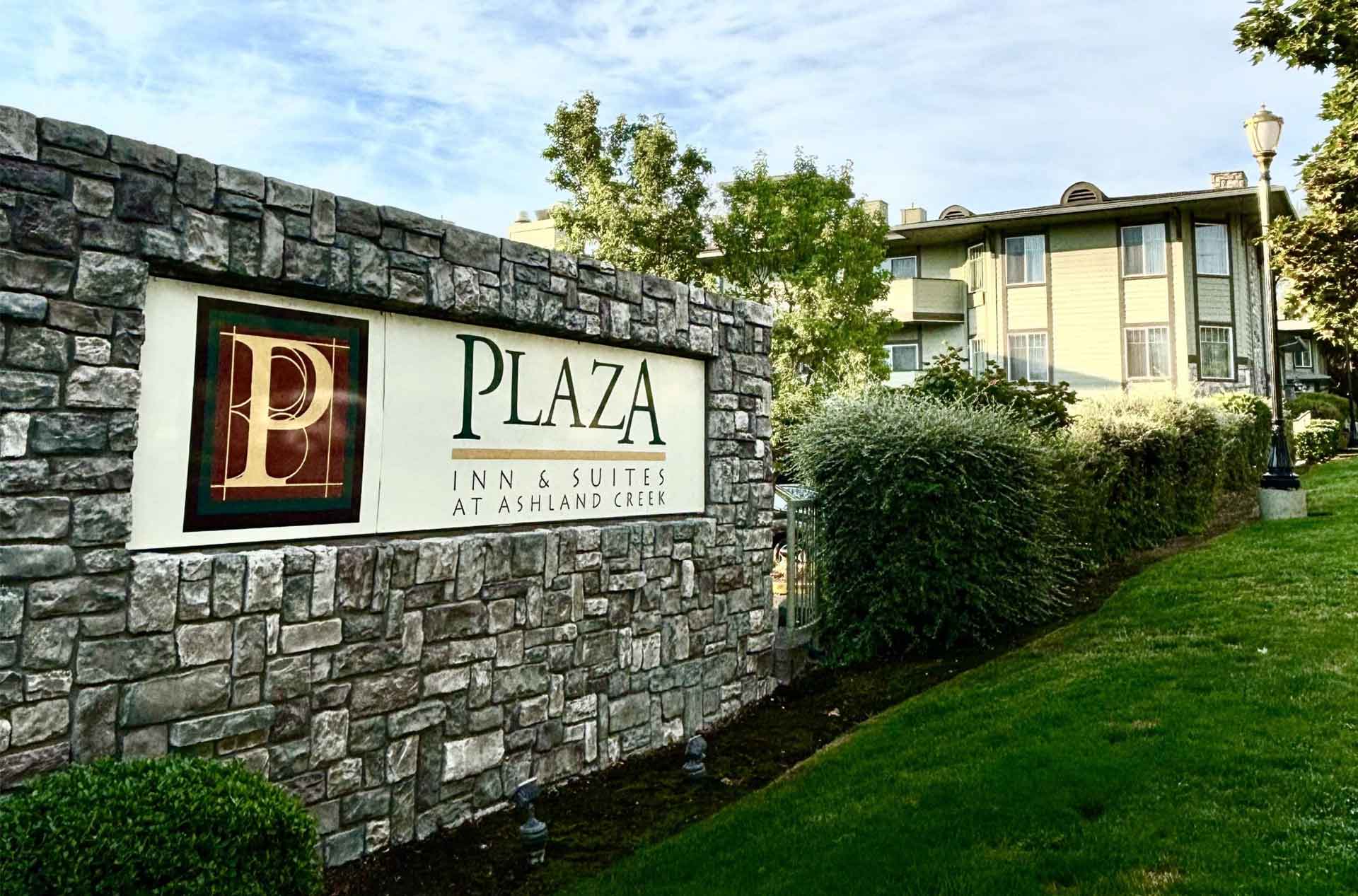 A stone sign for Plaza Inn and Suites at Ashland Creek, with a manicured green lawn and the building visible in the background on a clear day.