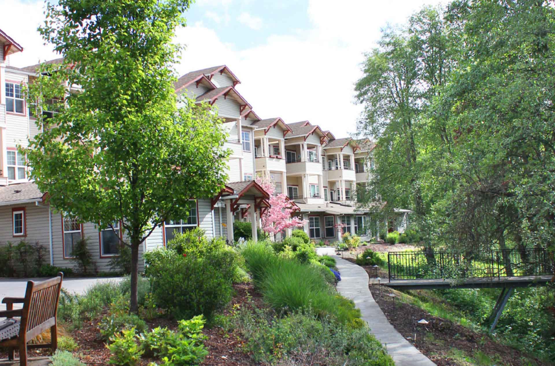 A retirement community building with red-trimmed gables, surrounded by landscaped gardens, trees, and a curved path leading to a small footbridge over a creek.