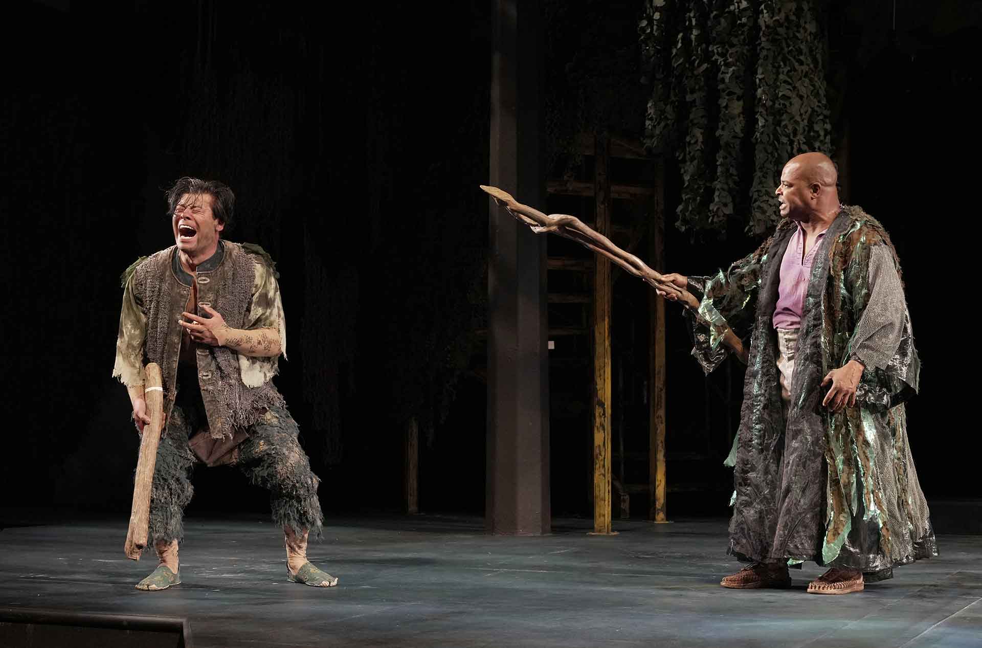 Man looking to the left laughing manically while a man on the right points a large wooden scepter.