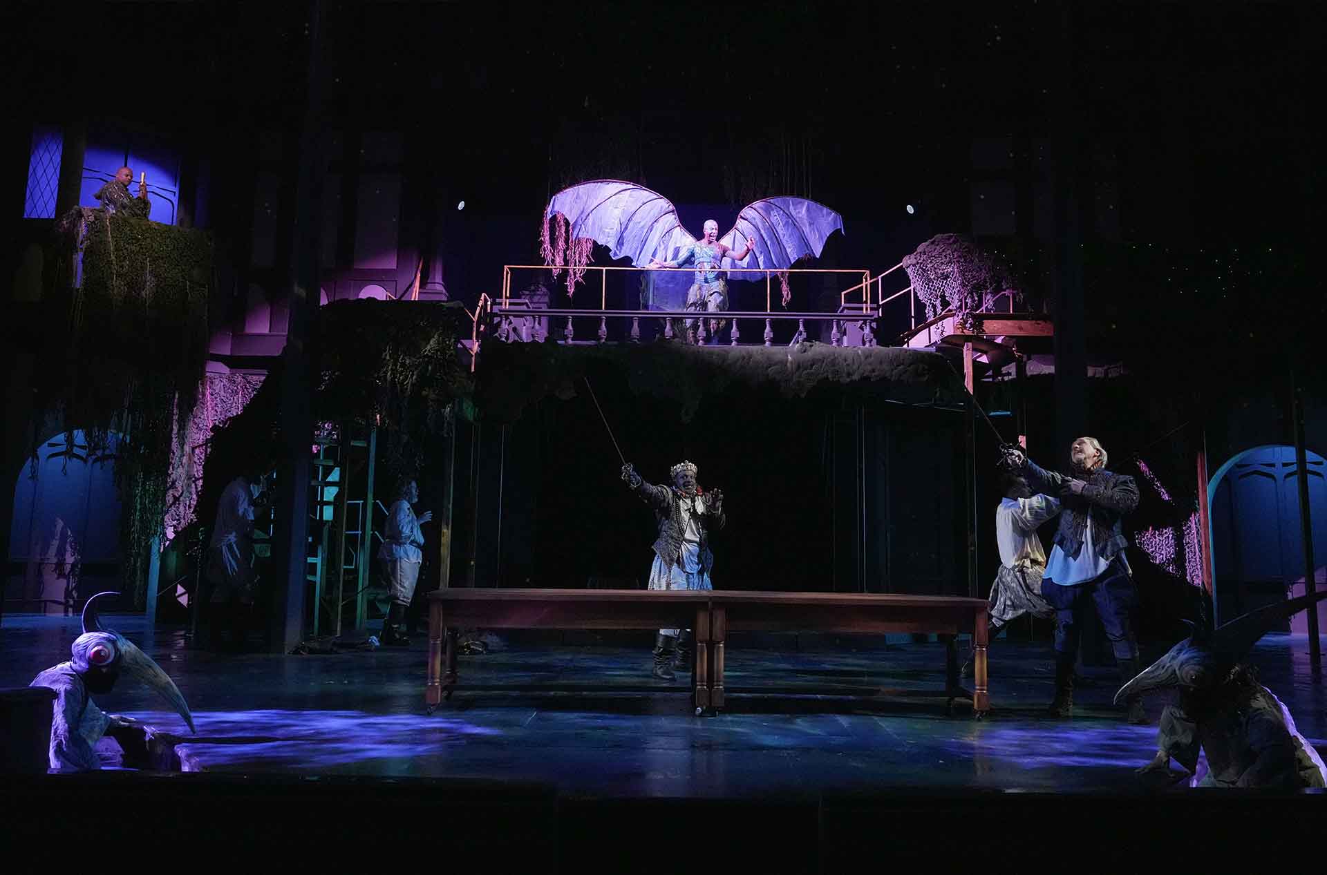 Wide view of stage, with people engaged in various activities and a winged creature at top.