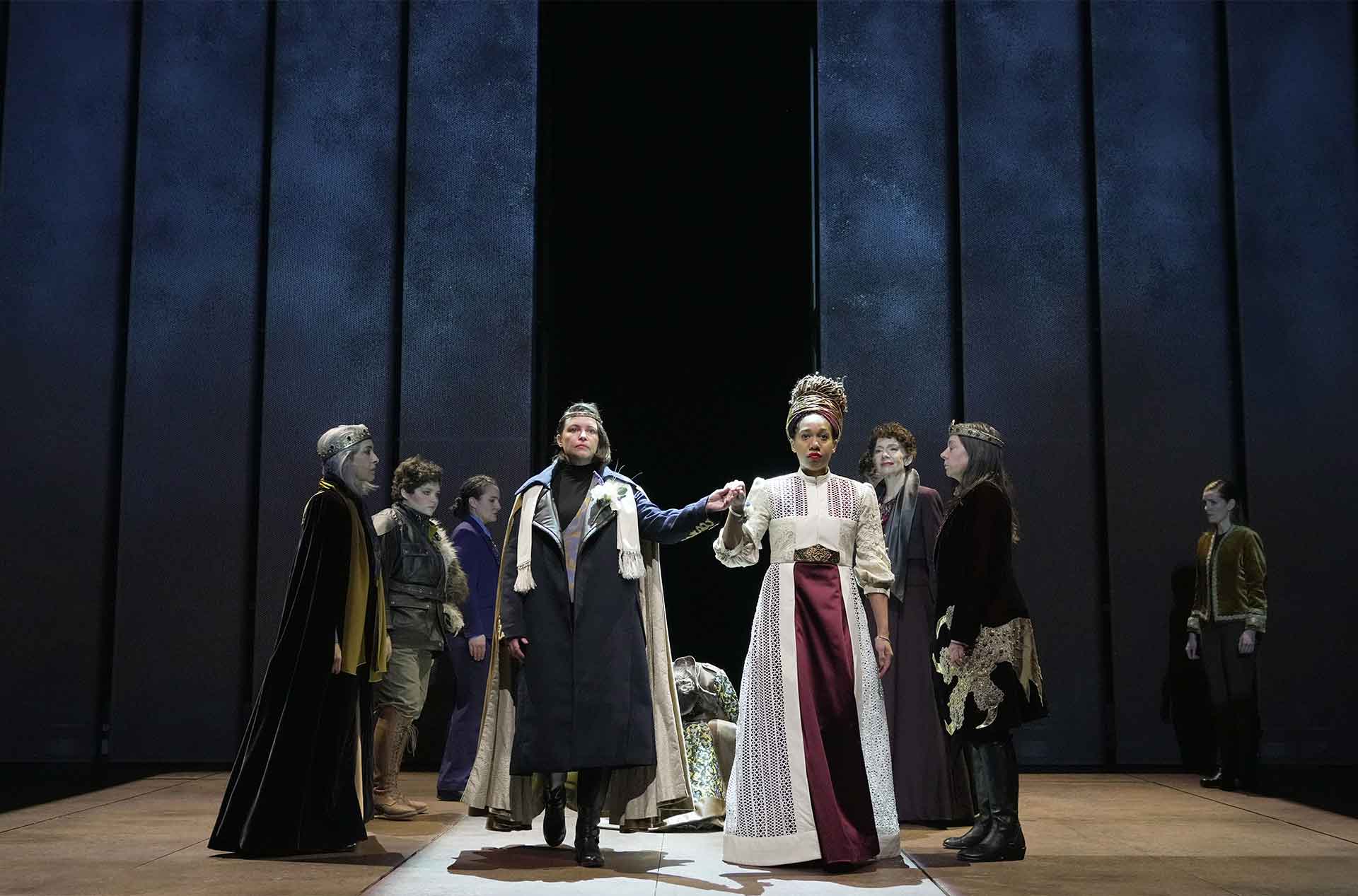 Two people holding hands and facing forward while two rows of people behind them face each other.