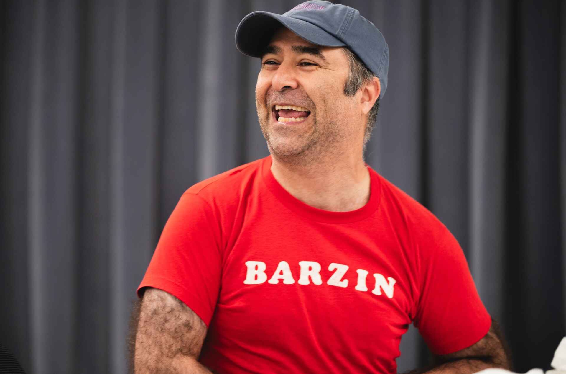 A  person wearing a red t-shirt with "BARZIN" printed on it and a dark blue baseball cap is sitting on a chair at a table. They seem engaged in conversation. The background features a curtain.