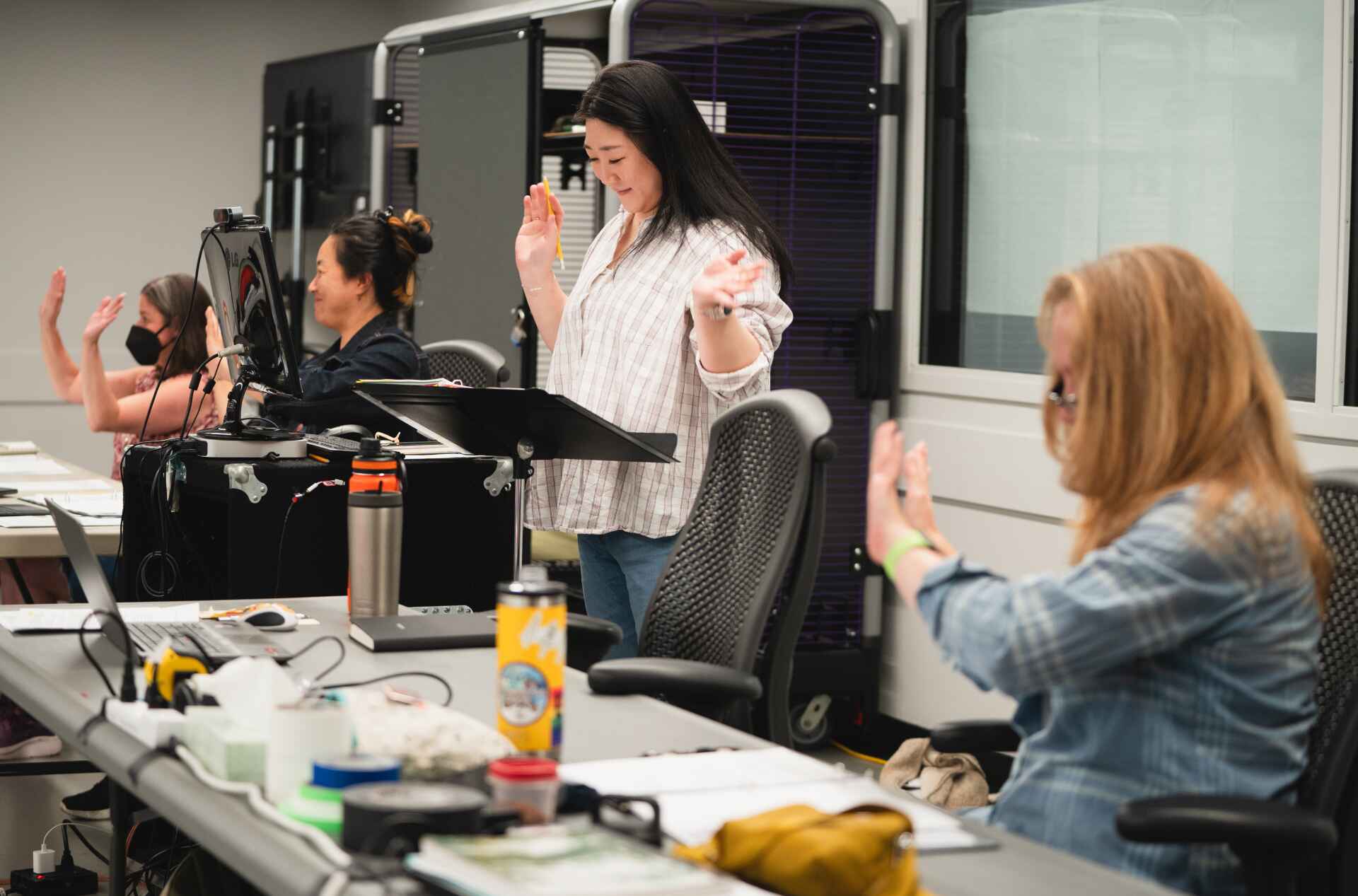 A group of individuals participate in rehearsal, with some raising their hands. They are seated at tables with various materials and a computer setup.
