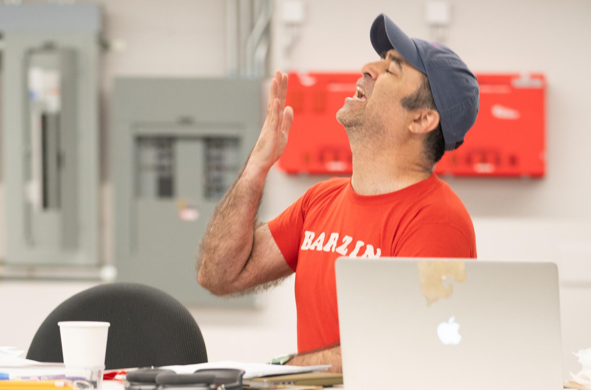 A person wearing a red shirt and hat sits at a table with a laptop, raising their head and hand in an expressive manner. Office materials are scattered on the table.