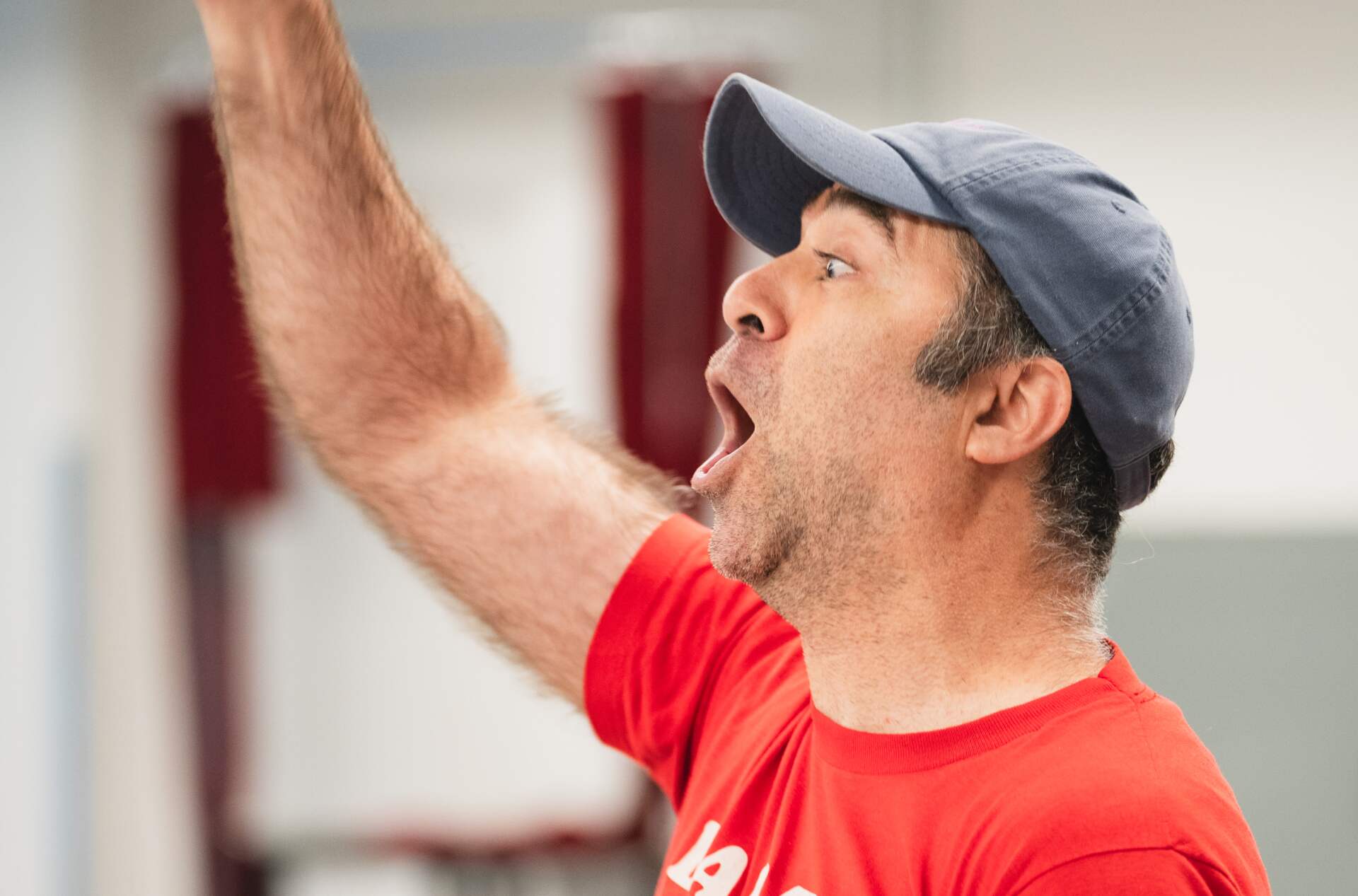 A person wearing a red t-shirt with white text and a blue cap is sitting and raising  Their right fist enthusiastically. Their mouth is open as if shouting or cheering.