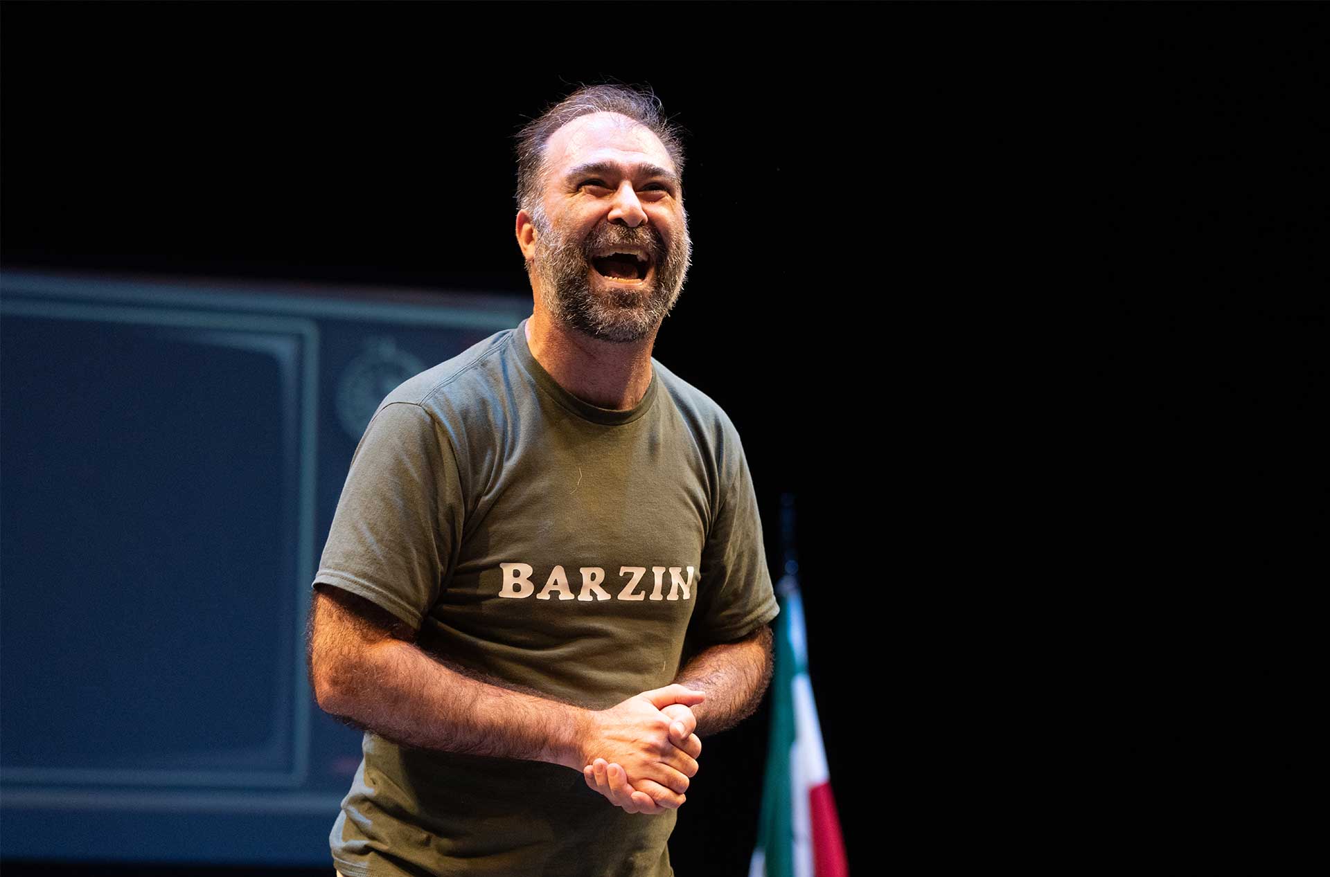 A person stands on stage smiling widely, wearing a shirt that reads "Barzin," with a flag in the background.