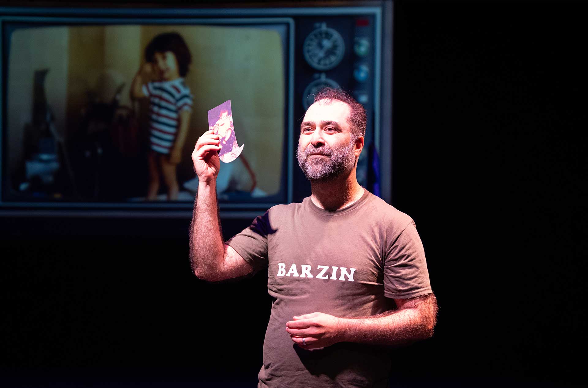 A person holds up a small photograph in one hand, standing in front of a projected image of a child on a vintage TV screen, while wearing a "Barzin" shirt.