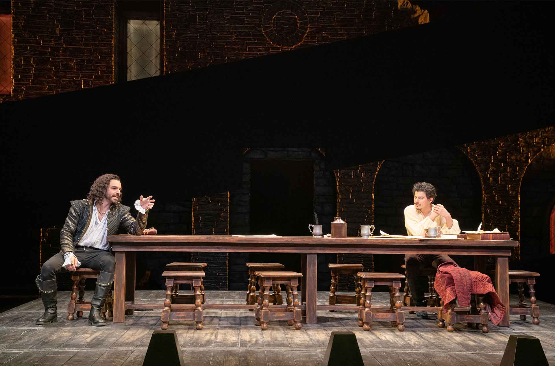 Two people in medieval-style outfits are seated at opposite ends of a long wooden table. The person on the left gestures with one hand, while the person on the right looks at them. The table has metal cups and a folded red cloth.