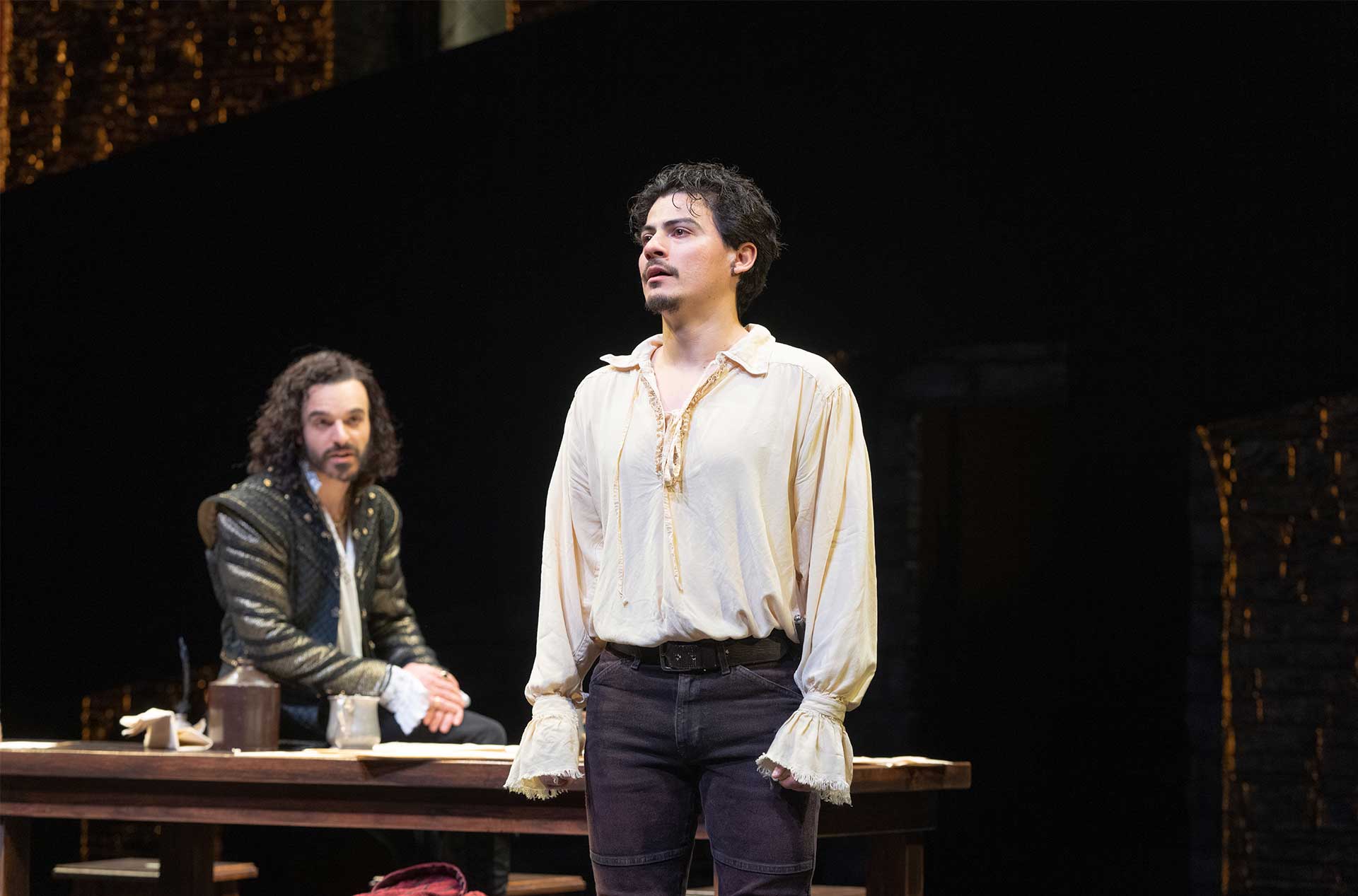 A person in a light-colored shirt stands in the foreground, looking into the distance. Another person with long curly hair and a dark outfit sits at a table in the background, watching the standing person.