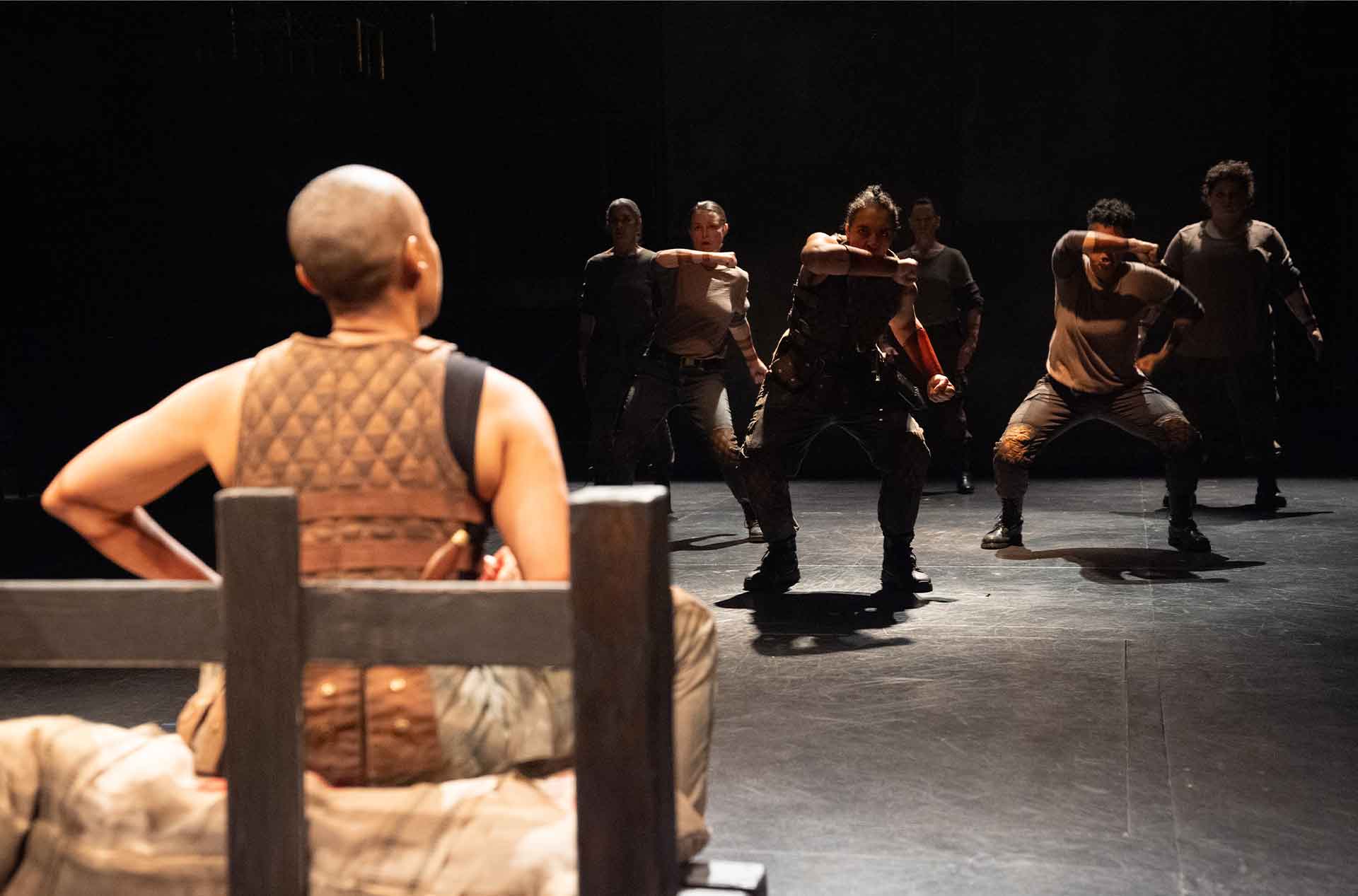 A person with a shaved head sits facing a group who are positioned in intense dynamic poses as if preparing for a confrontation. The dim lighting and shadowy background add to the scene's tension.