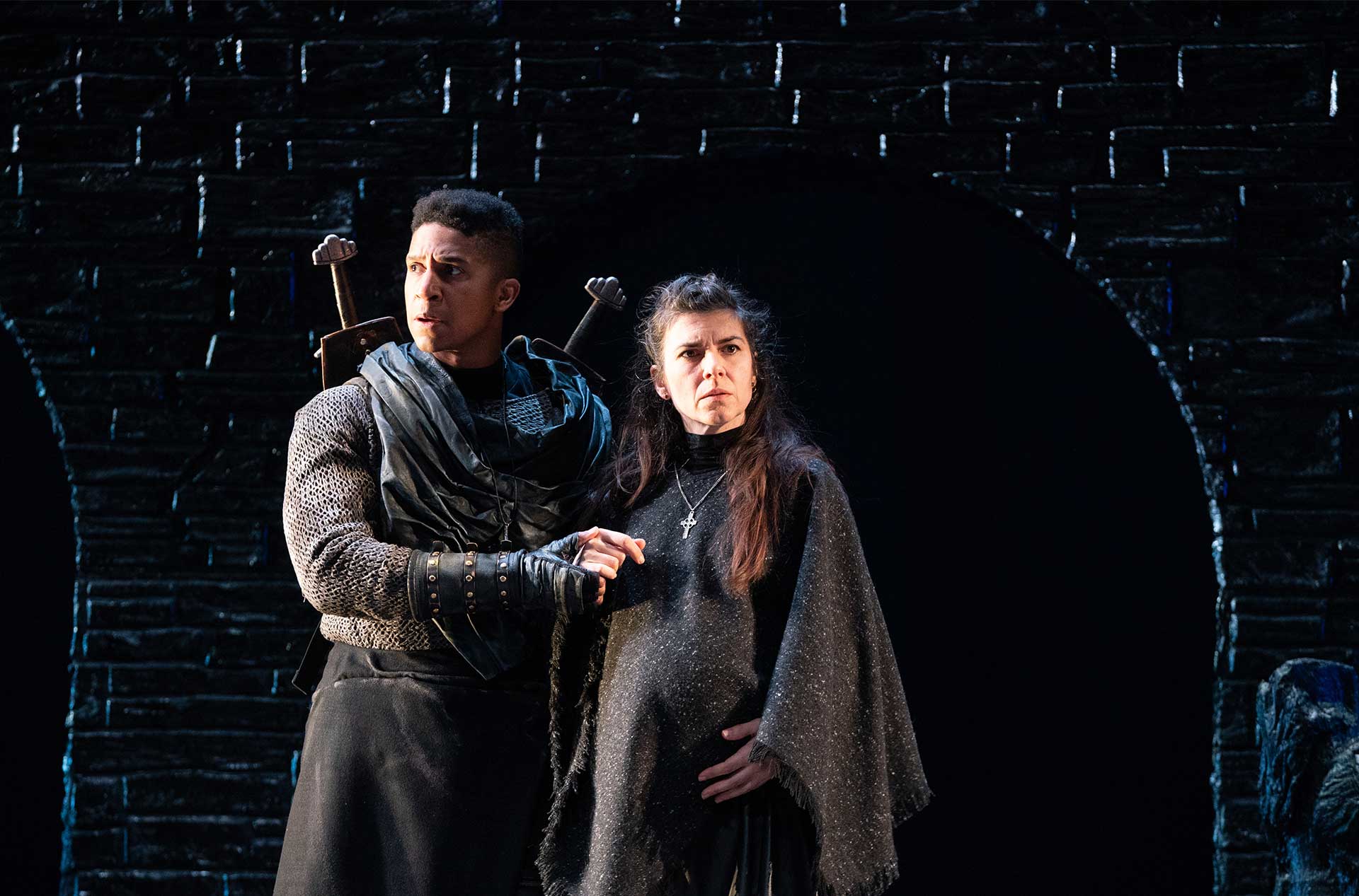 A person with crossed swords on their back supportively holding the arm of a pregnant person. Both are wearing medieval-style costumes.