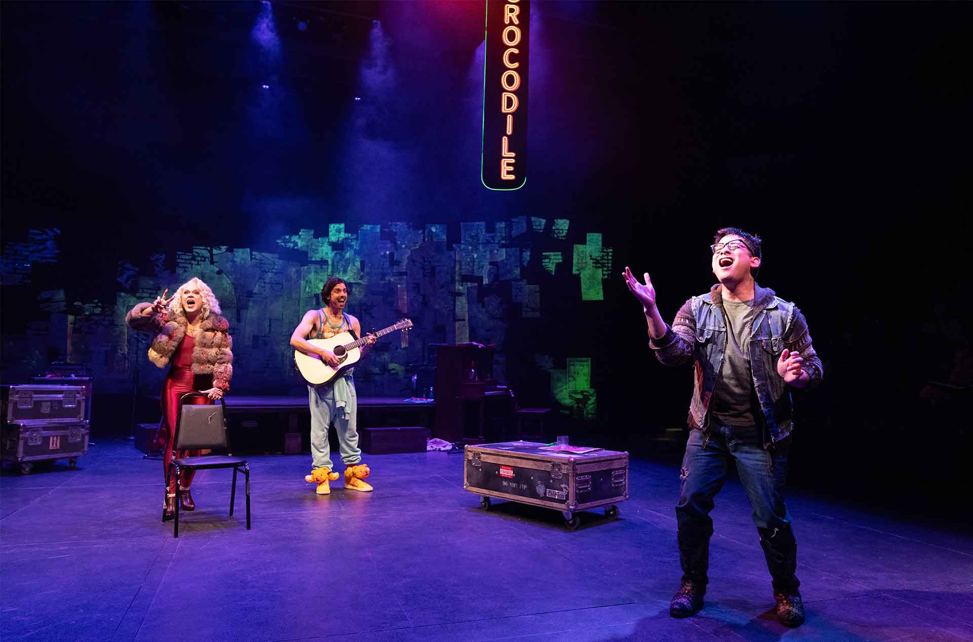Three people are on stage with a "Crocodile" sign overhead. On the left, a person in a fur coat and red outfit stands near a chair. In the center, a person in a tank top and teddy bear shoes plays a guitar. On the right, a person in a denim jacket sings with one hand raised.