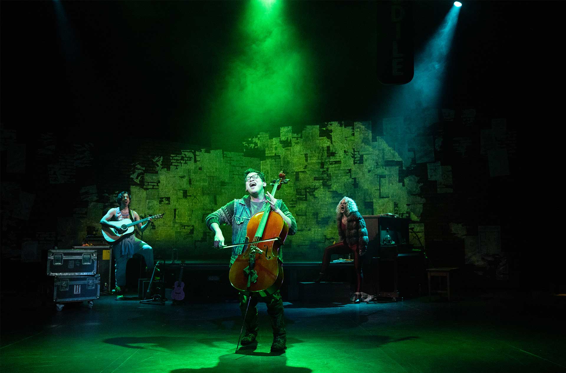 Green light shining down on a person playing the cello with two others in the background.