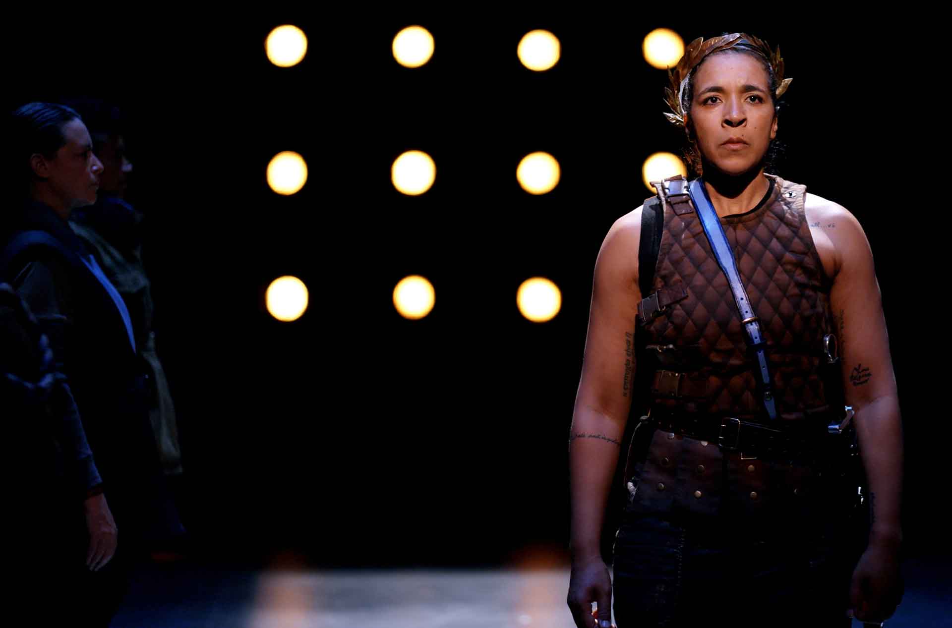 A person stands on stage wearing a quilted vest with straps, looking resolutely forward. The background is dark with rows of illuminated circular lights, creating a dramatic atmosphere. Other figures are partially visible in the background, also facing forward.