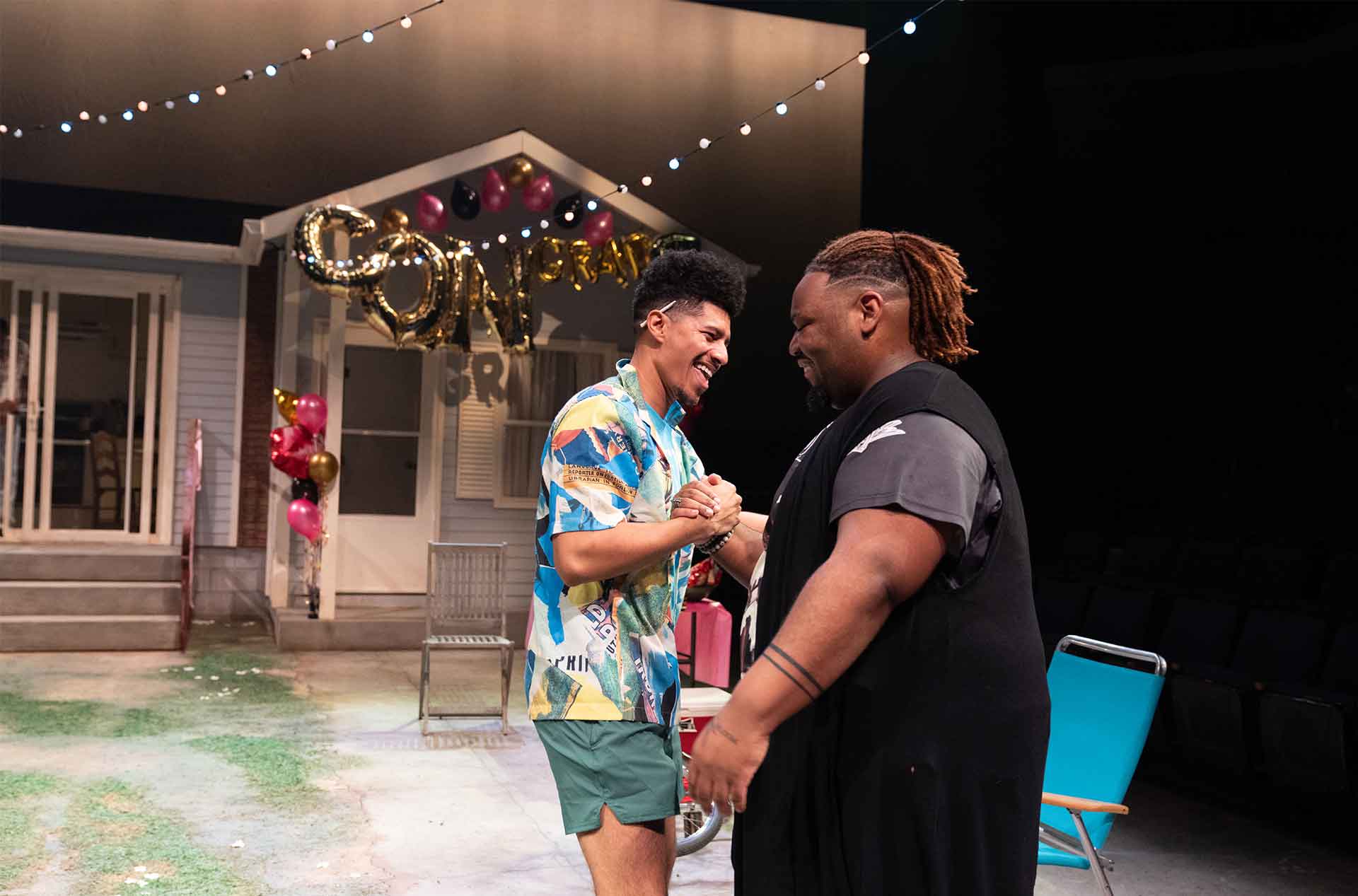 Two people laugh and shake hands near a house decorated with balloons and a “Congrats Grad” sign.