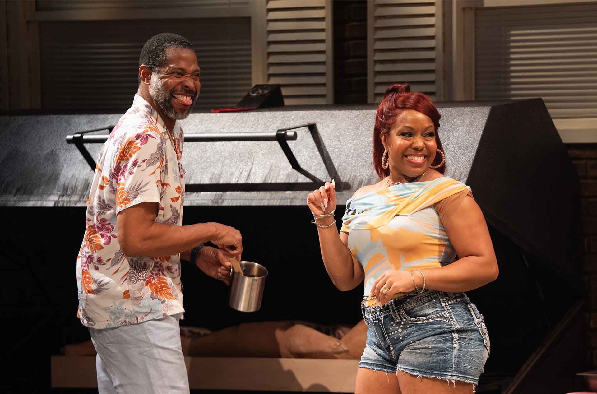 Two people stand in front of a grill, smiling and appearing mid-laugh during a lighthearted moment.