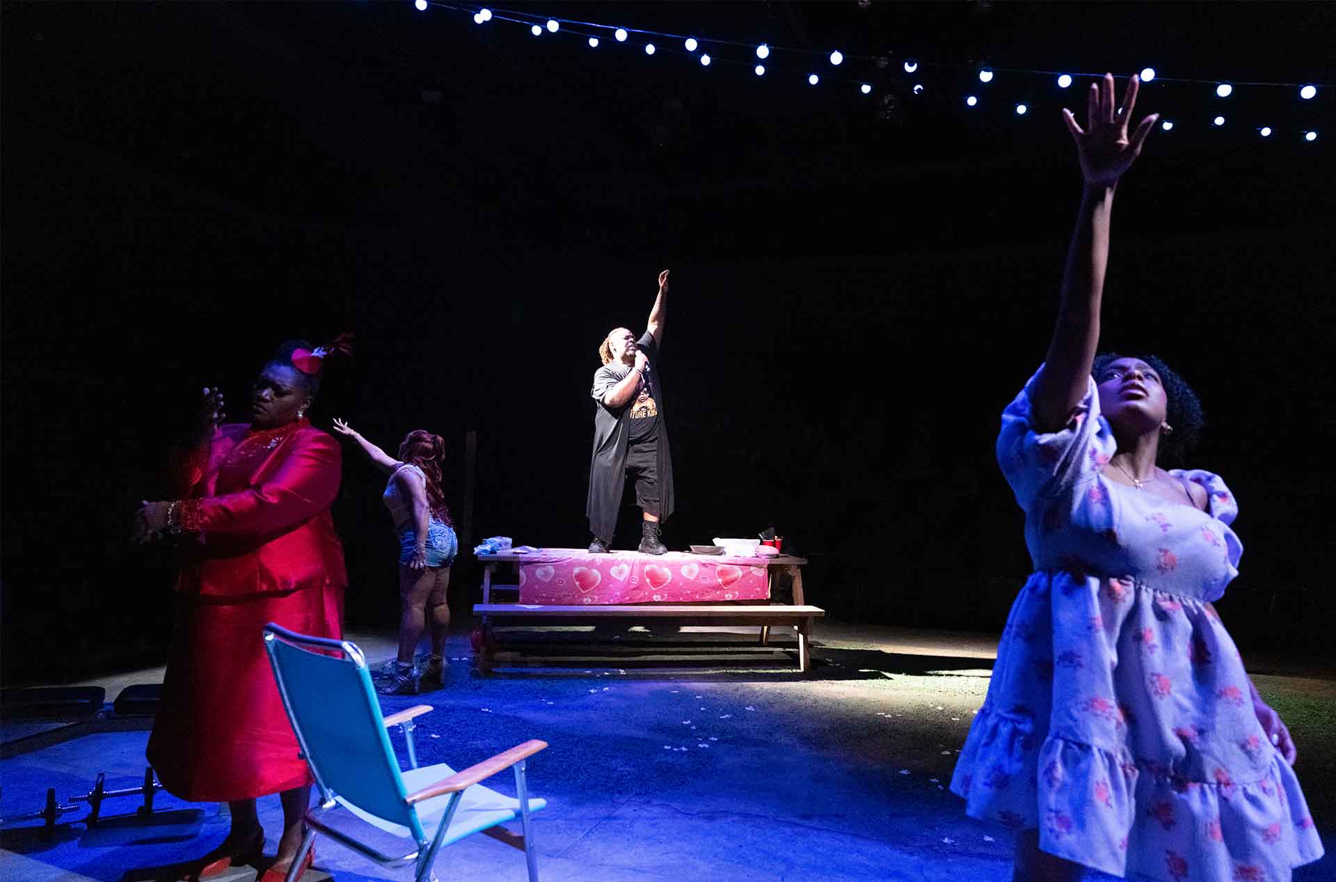 A group of people strikes expressive poses under dramatic blue lighting, one standing on a picnic table with an arm raised.