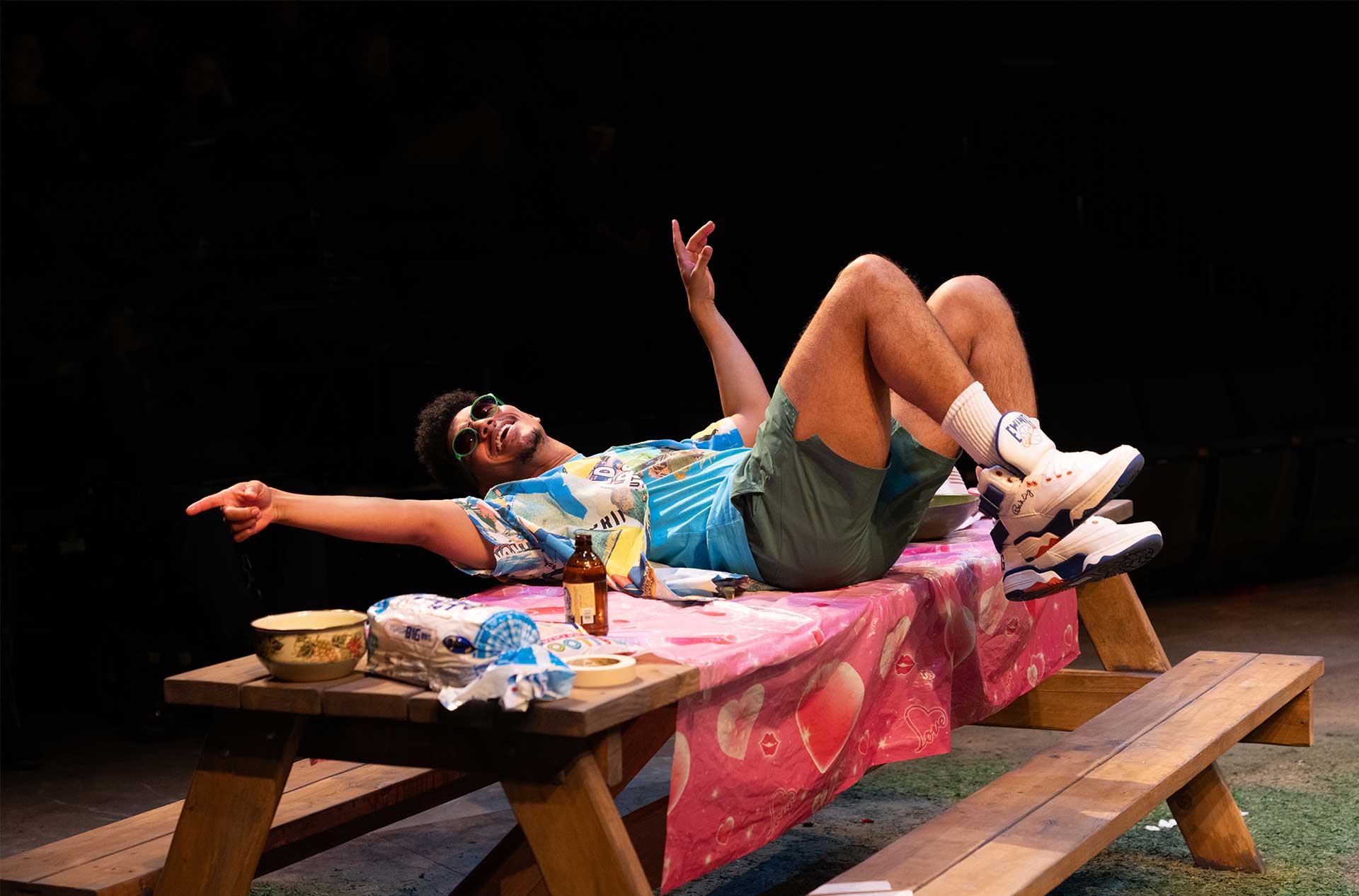 A person in sunglasses and a colorful shirt lounges across a picnic table, pointing and smiling under party lights.