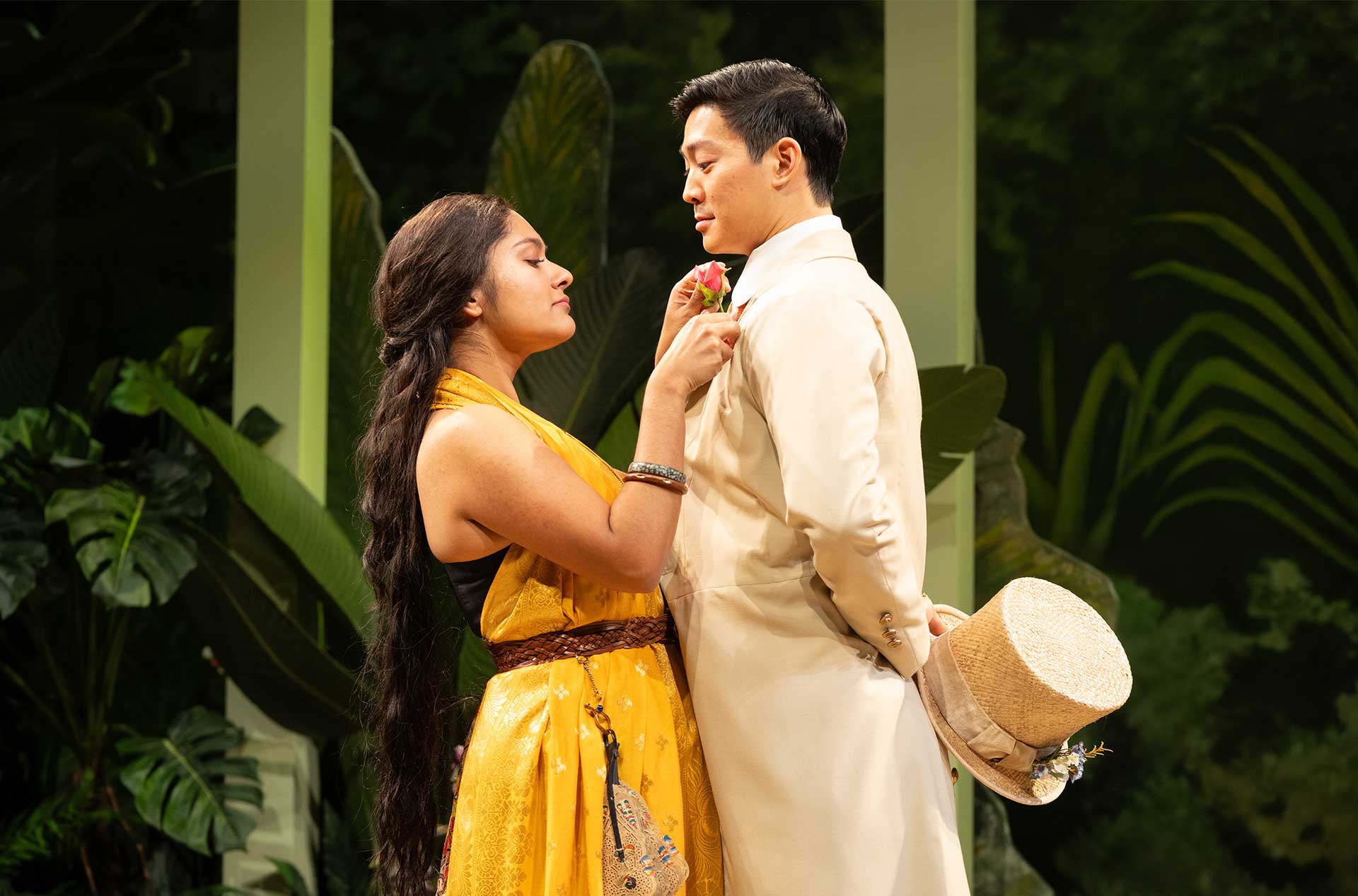 A person in a yellow outfit pins a flower to another’s lapel in a lush outdoor setting, both gazing at each other.