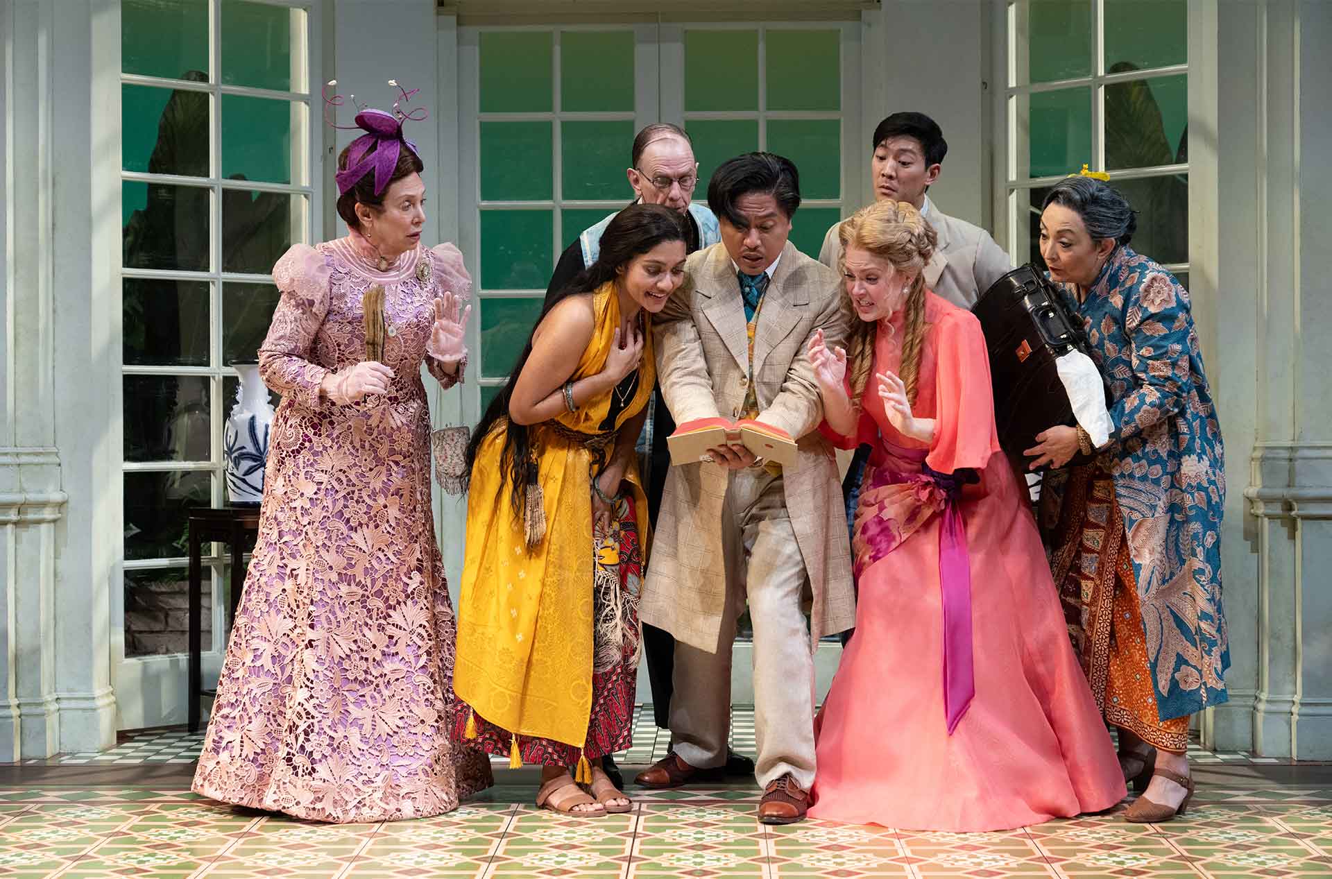 A group of actors in vibrant period costumes huddle around a book, displaying expressions of shock, excitement, and curiosity.