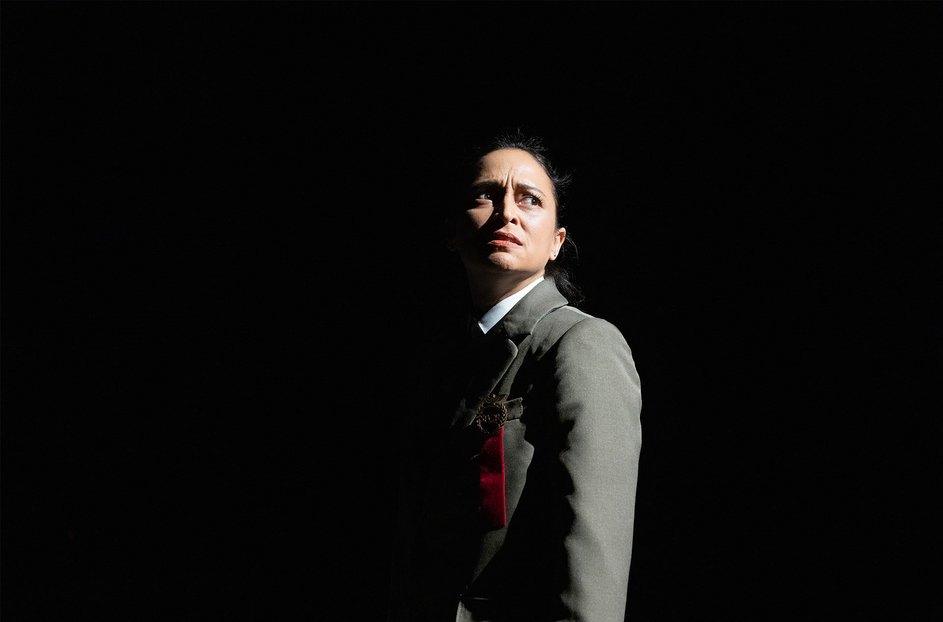 A character in a dark suit stands alone under dramatic lighting, their face partially illuminated, looking upward with a tense expression.