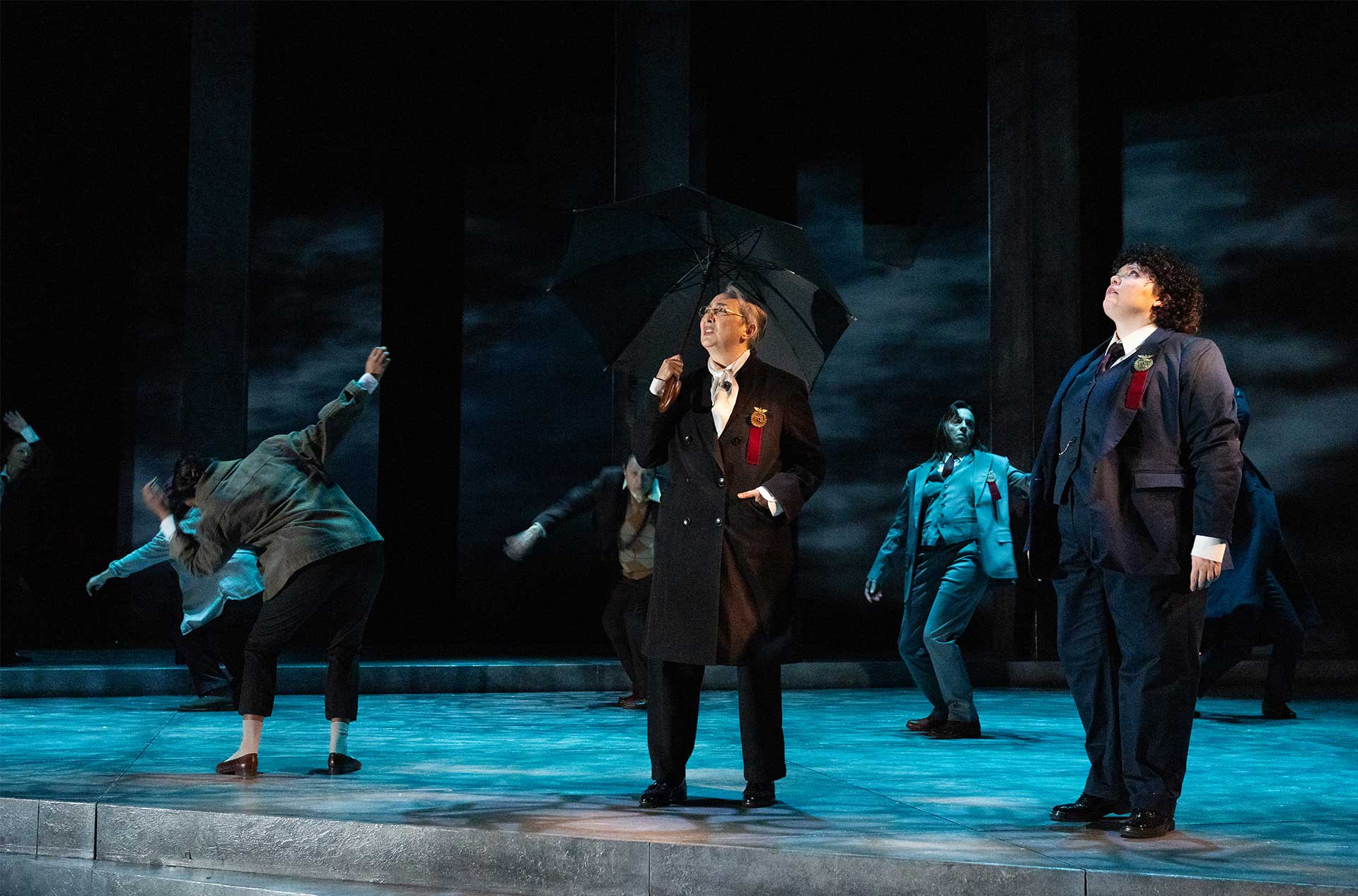 A figure in a long black coat holds an umbrella and looks upward, surrounded by others in suits reacting dynamically under a stormy, blue-lit stage.
