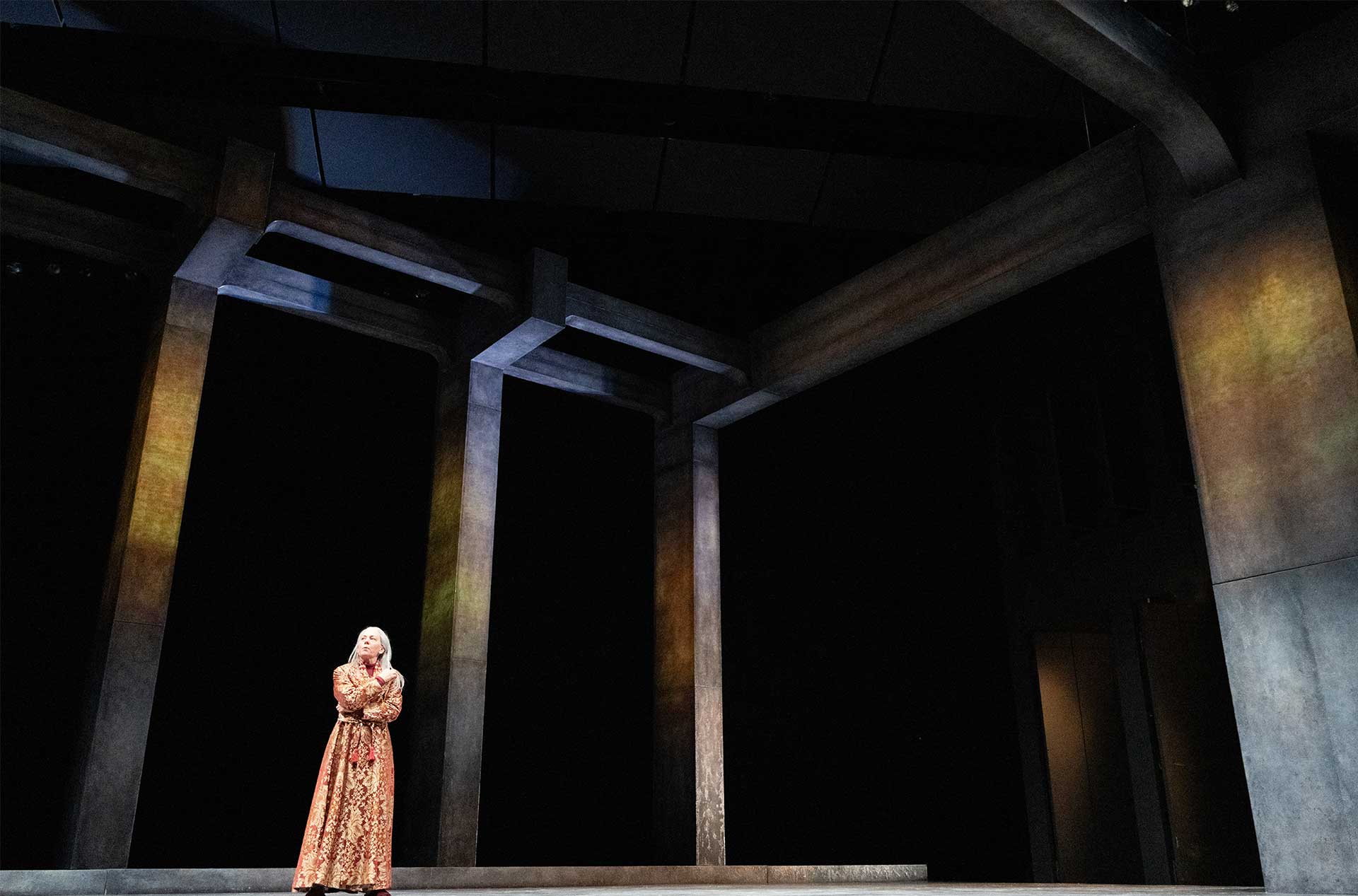 A lone figure in an ornate, gold-patterned robe stands on a vast, shadowed stage, framed by towering, angled columns.