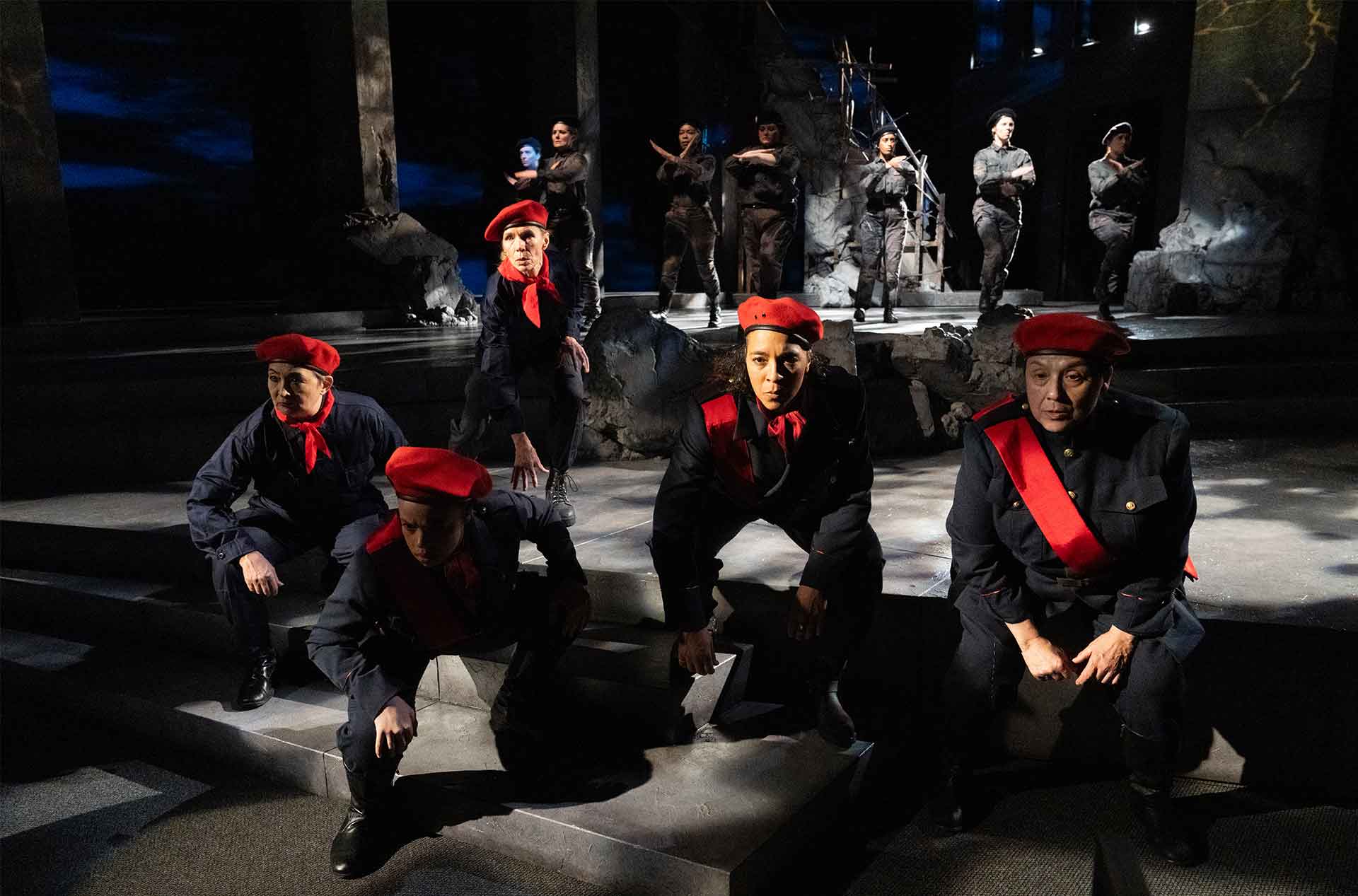A group of performers in black military uniforms with red berets crouch in formation, while others in darker attire stand in the background against a ruined, stone-like set.