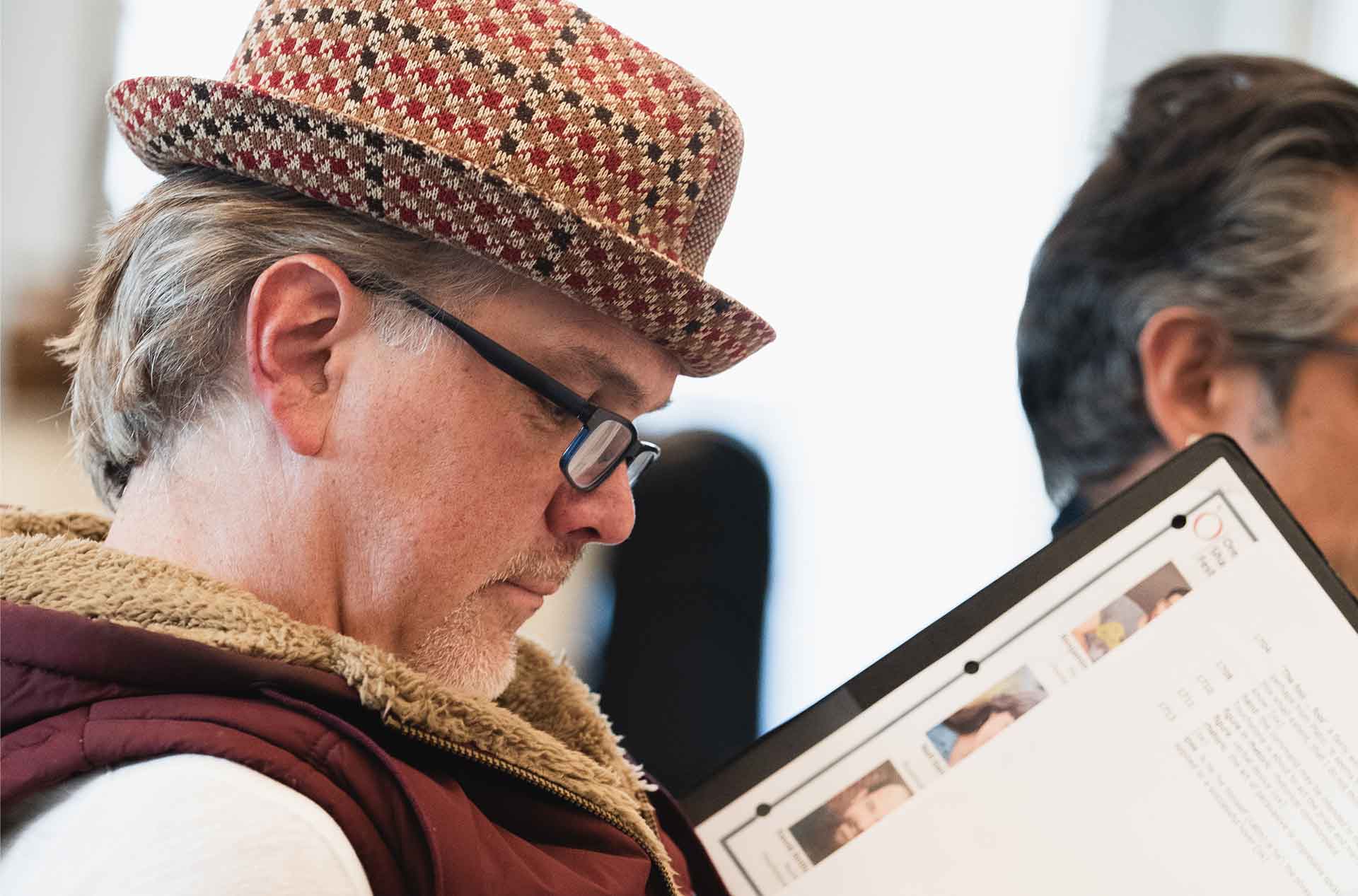 A person wearing a patterned hat and glasses closely examines a binder with headshots and text. Another person is slightly out of focus in the background.