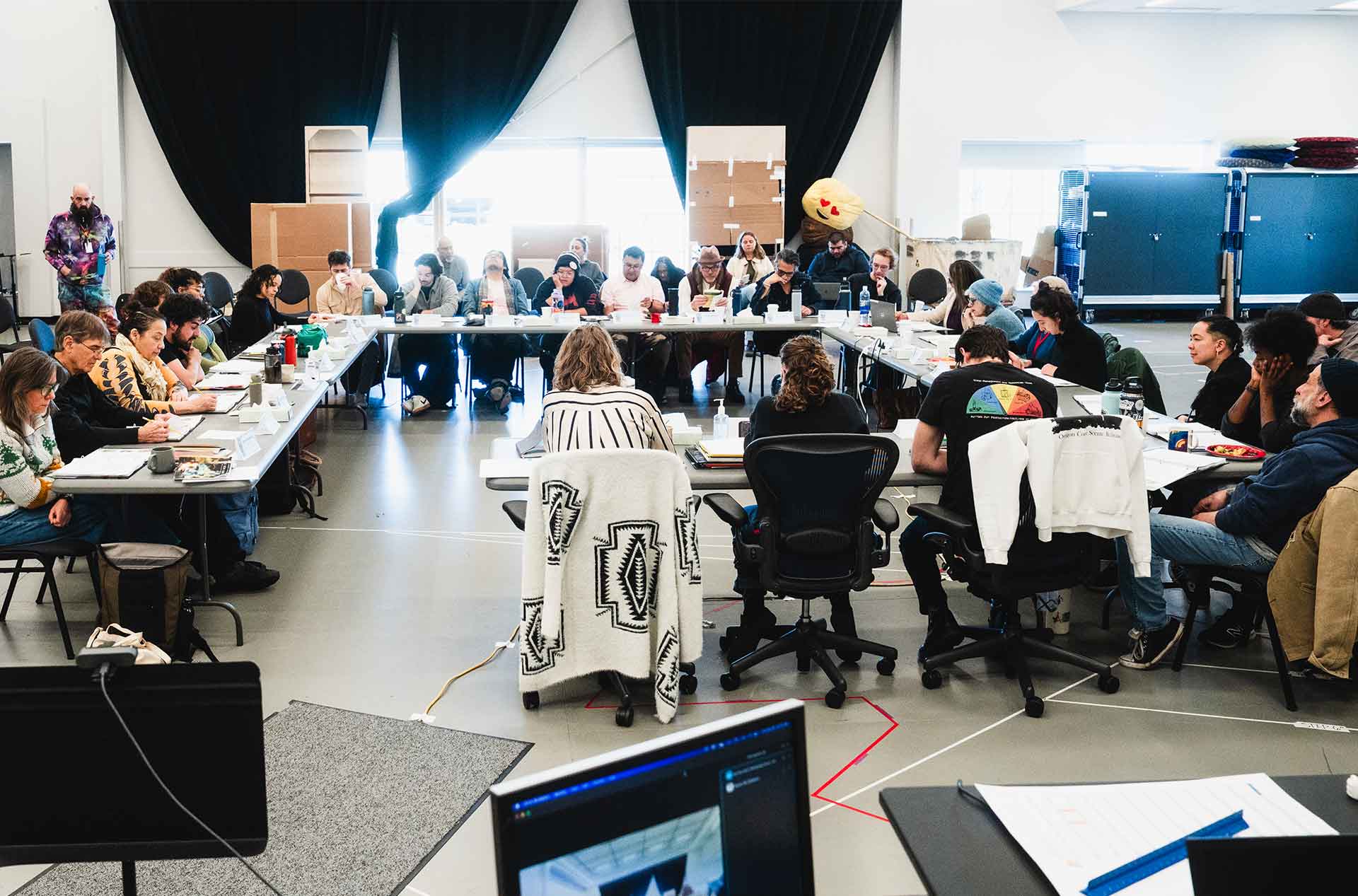 A wide view of a reheasal room with a long U-shaped table, where many people are engaged in discussion or taking notes. Large boxes and a plush emoji decoration are in the background.