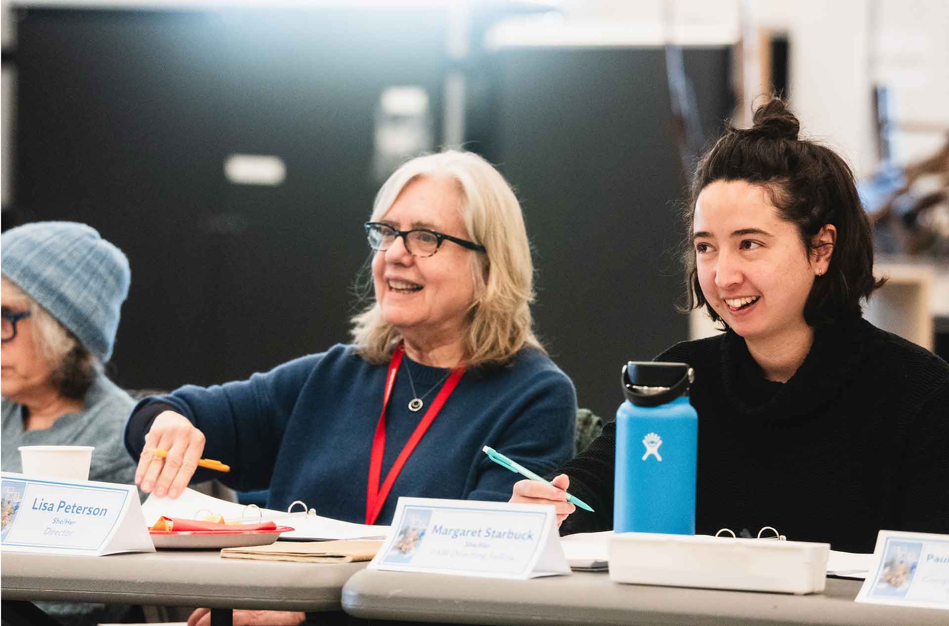 Two people sit at a table during a discussion, both smiling. One person wears glasses and a lanyard, while the other holds a pen.