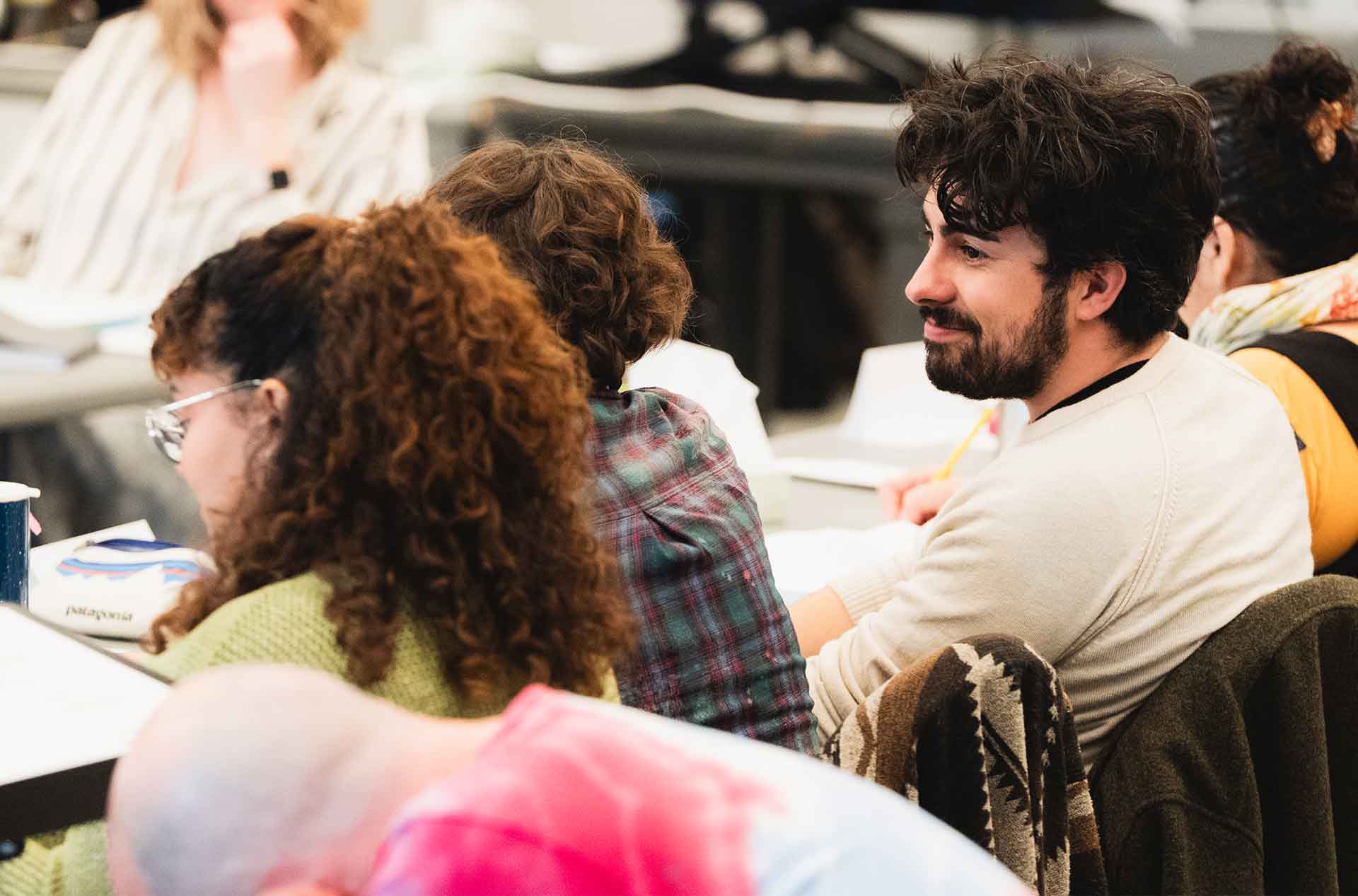 A person in a cream-colored sweater listens with a slight smile during a discussion. Others nearby are taking notes or talking.
