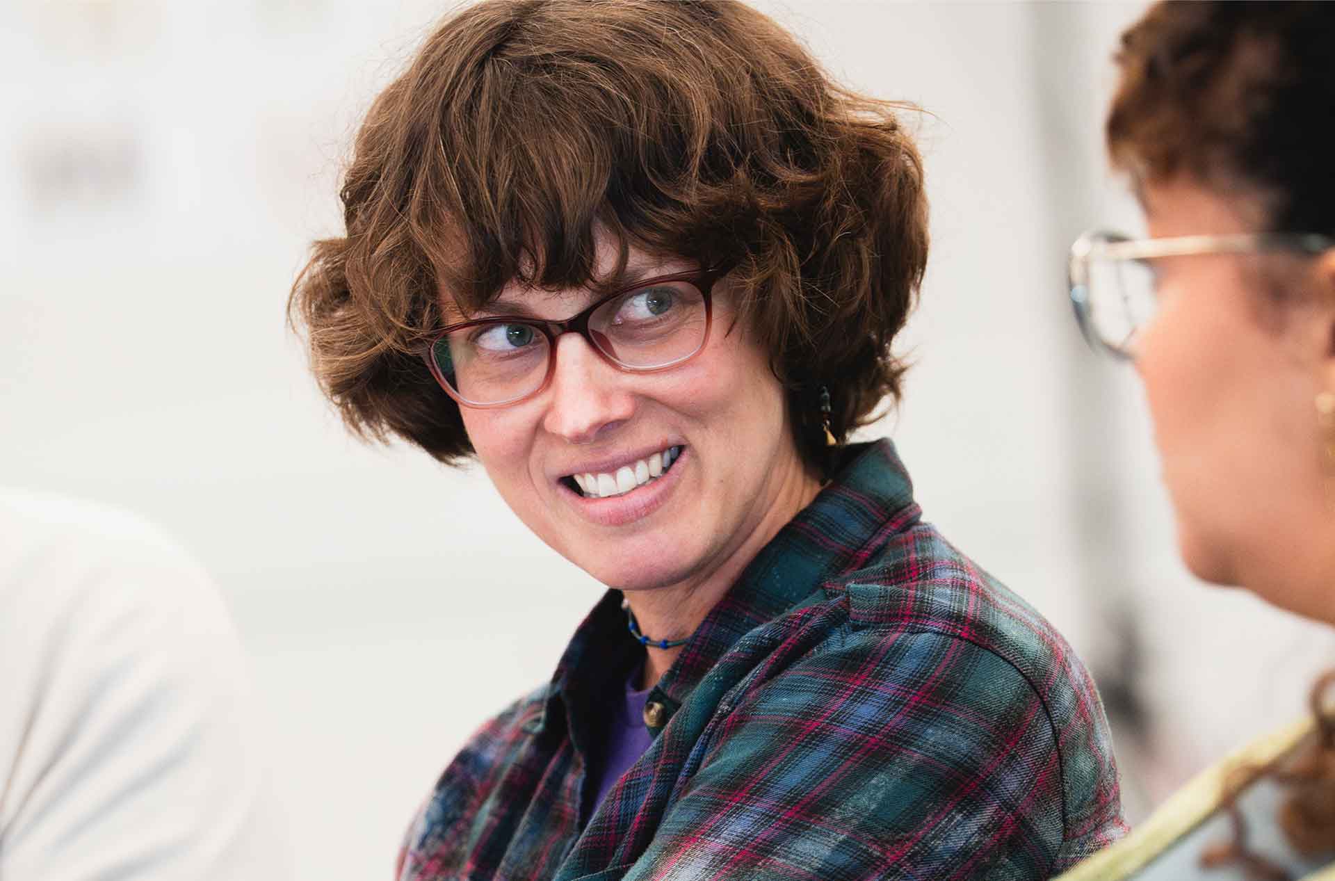 A person in a plaid shirt and glasses smiles while talking to someone nearby. They appear engaged in conversation.