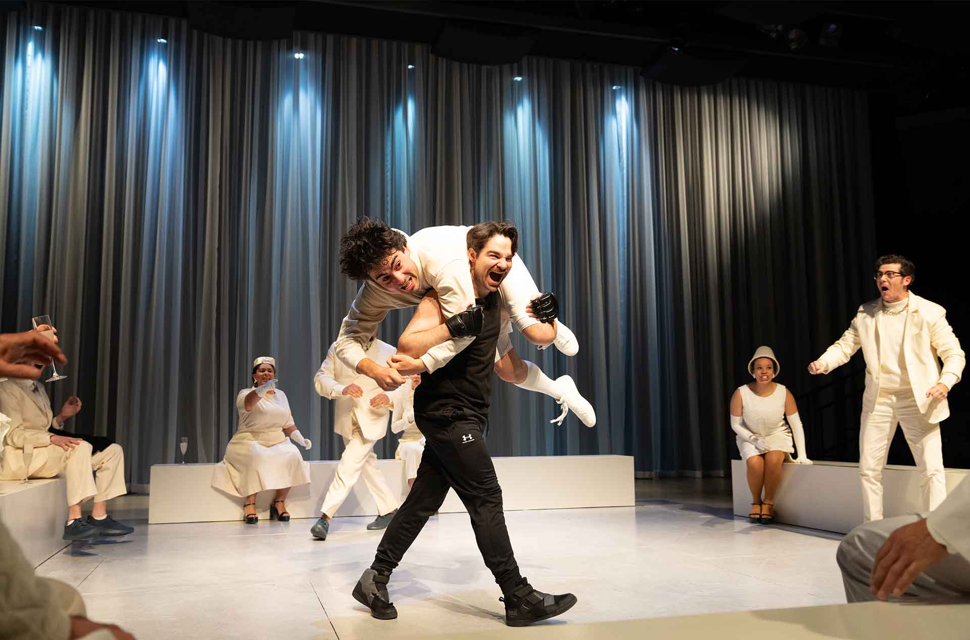 A high-energy scene where a performer in black carries another in white across their shoulders as others in matching white outfits react with surprise and excitement.