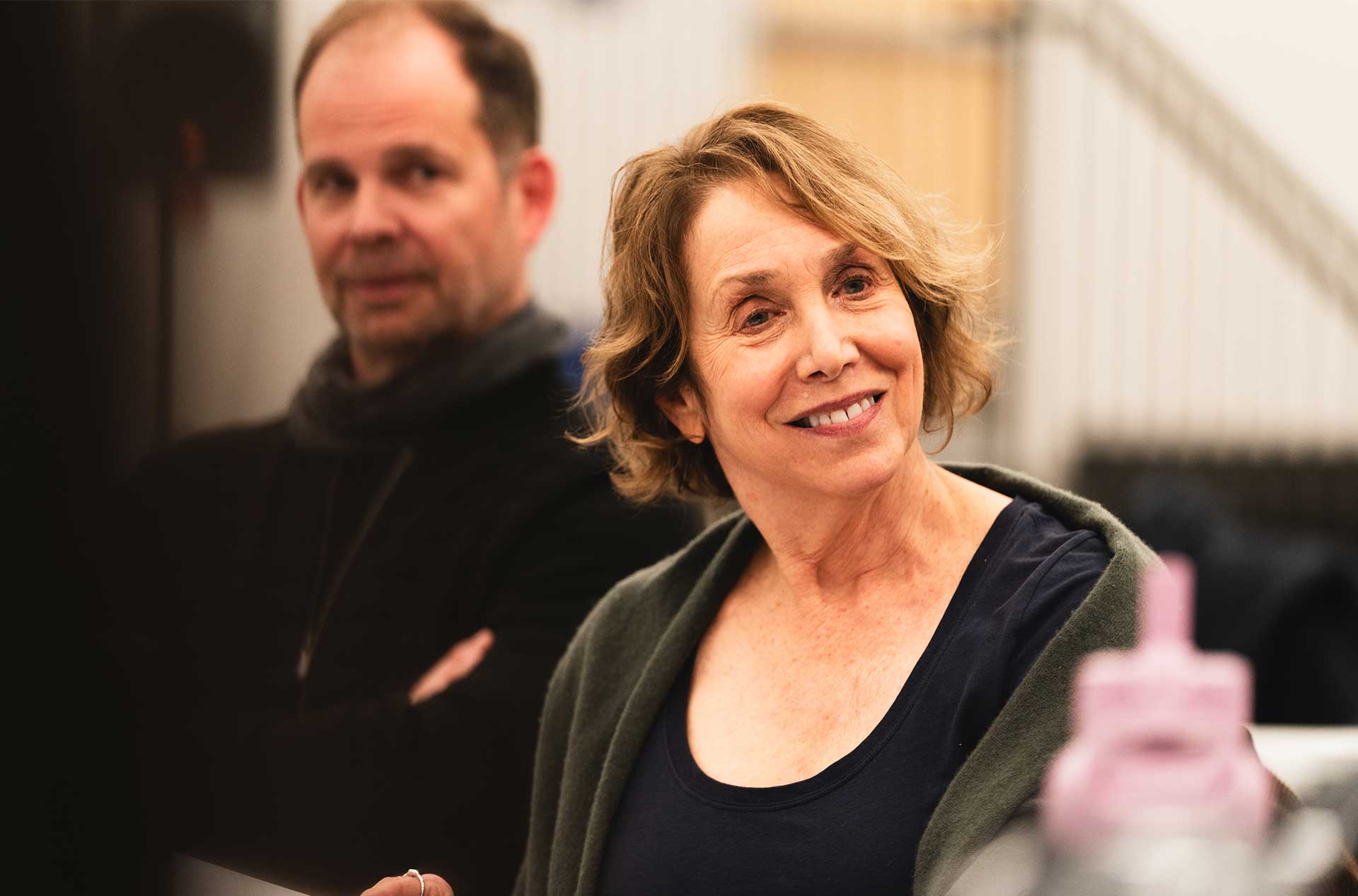 A person with short hair smiles warmly while seated at a table. A second person in the background looks on, appearing thoughtful.