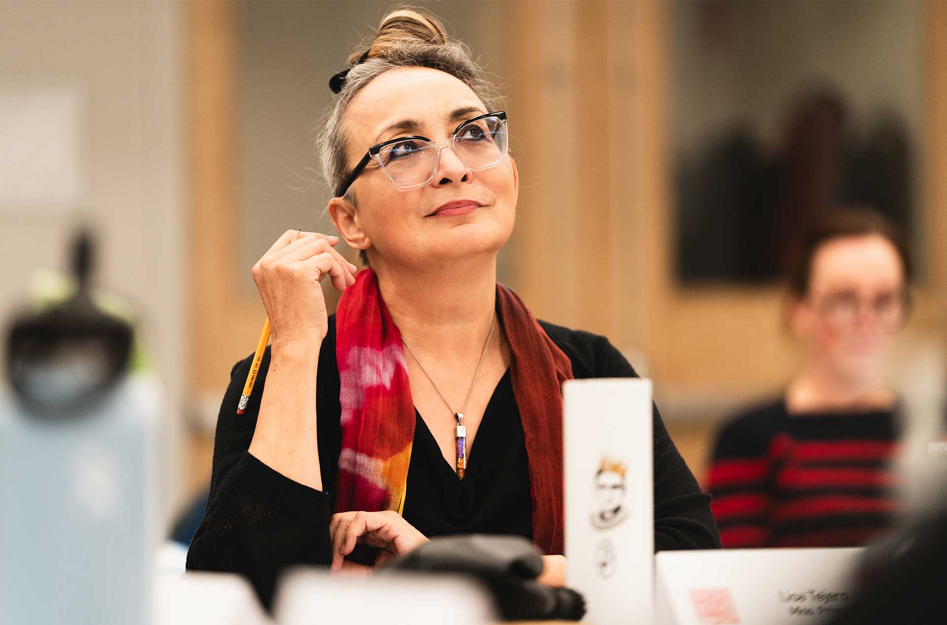 A person wearing glasses and a colorful scarf sits at a table, holding a pencil and looking thoughtful. Papers and nameplates are visible in the foreground.