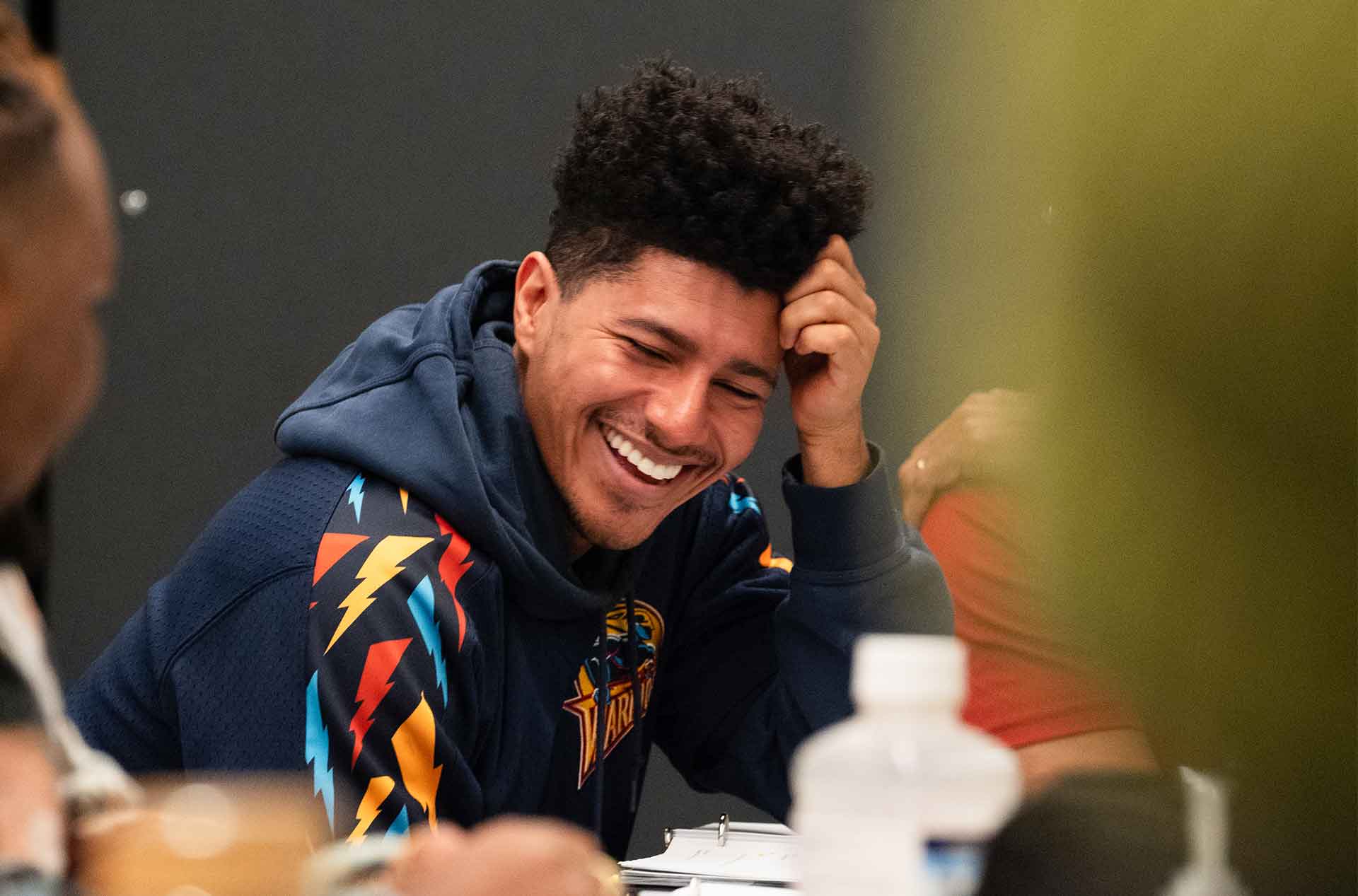 A person in a navy hoodie with a colorful design laughs while seated at a table. Their hand rests on their head, and their expression conveys amusement.