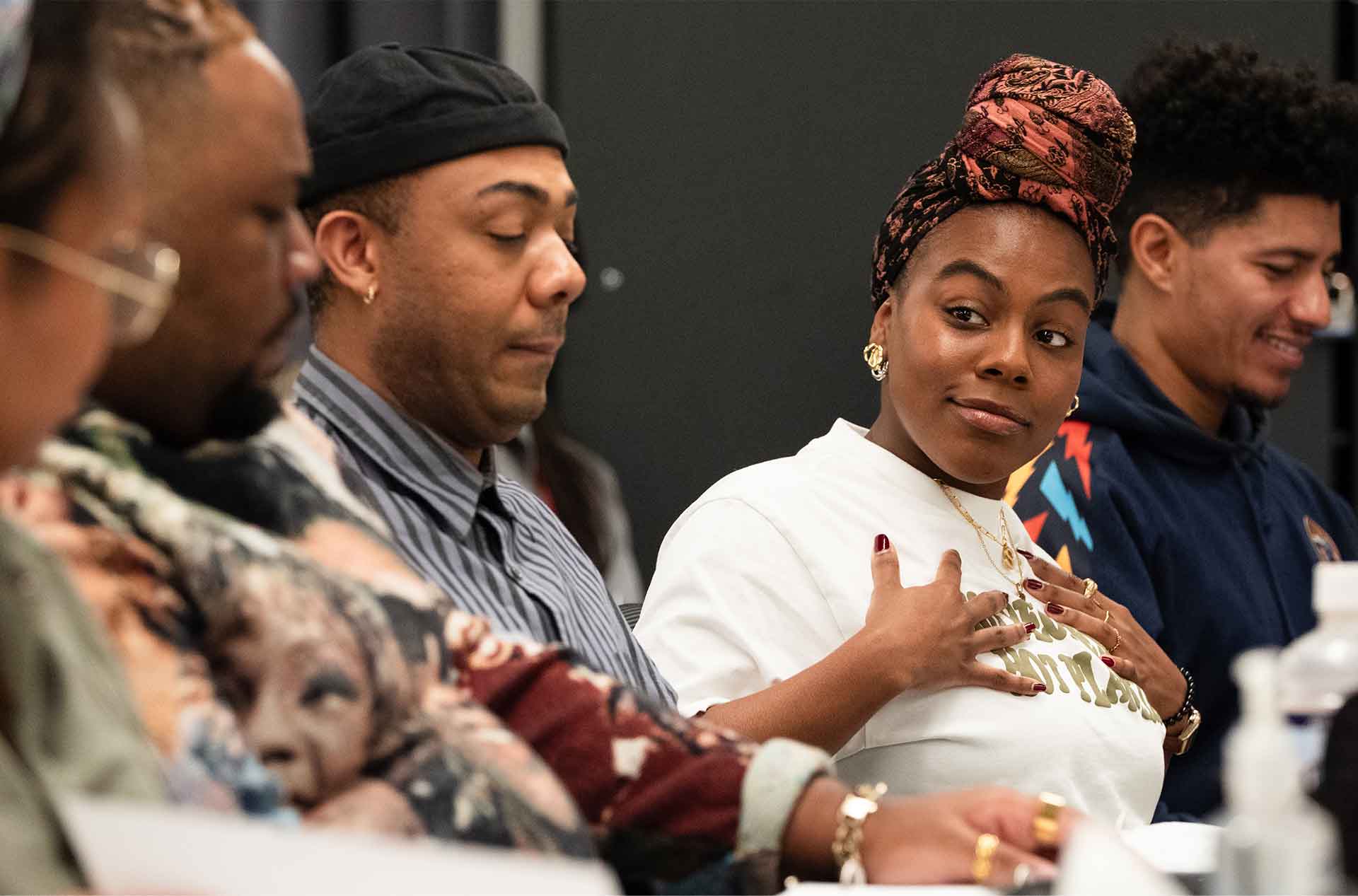 A group of people seated at a table in discussion. One individual with a headwrap and jewelry gestures toward their chest while looking at another person, who appears to be concentrating.