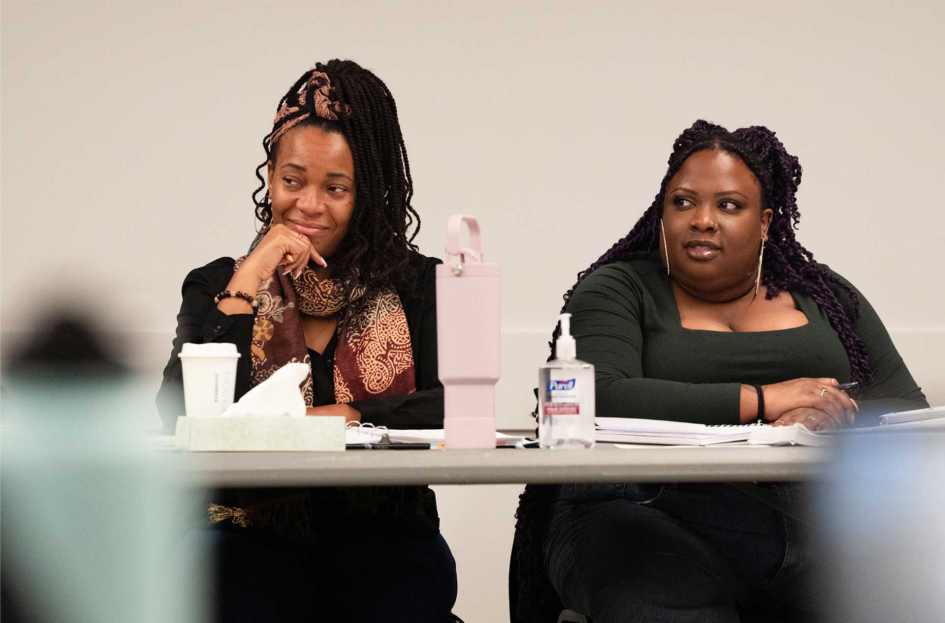 Two people sit at a table, reacting to something off-camera. One, wearing a scarf, rests their chin on their hand with a smirk, while the other, in a green top, looks on with a neutral expression.