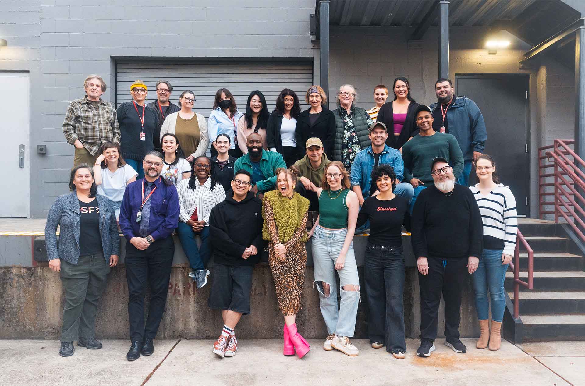 A large group of cast and crew members from "Into the Woods" pose outside a building, smiling at the camera on a loading dock.