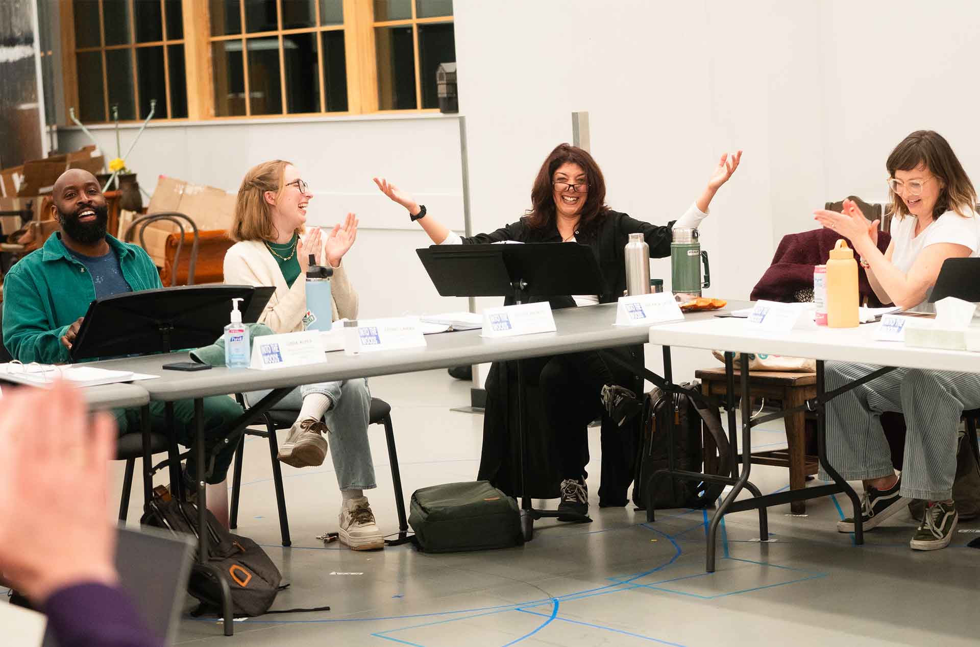 A group of people seated at a table laughing and clapping during a lively rehearsal session, with nameplates and water bottles in front of them.