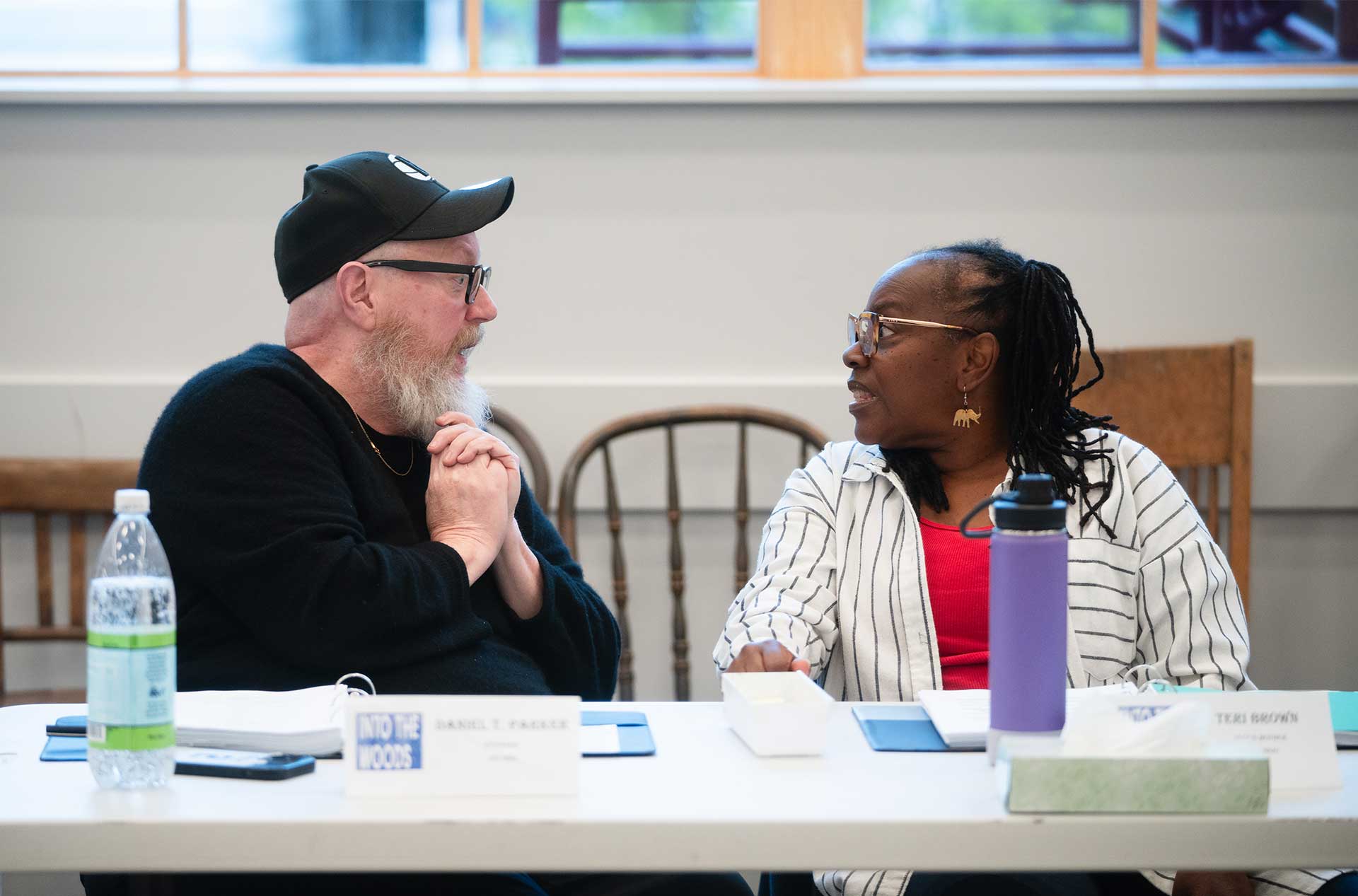 Two people sit at a table in conversation, one with a long white beard and black cap, the other wearing striped clothing and glasses. A nameplate reading “Into the Woods” is visible on the table.