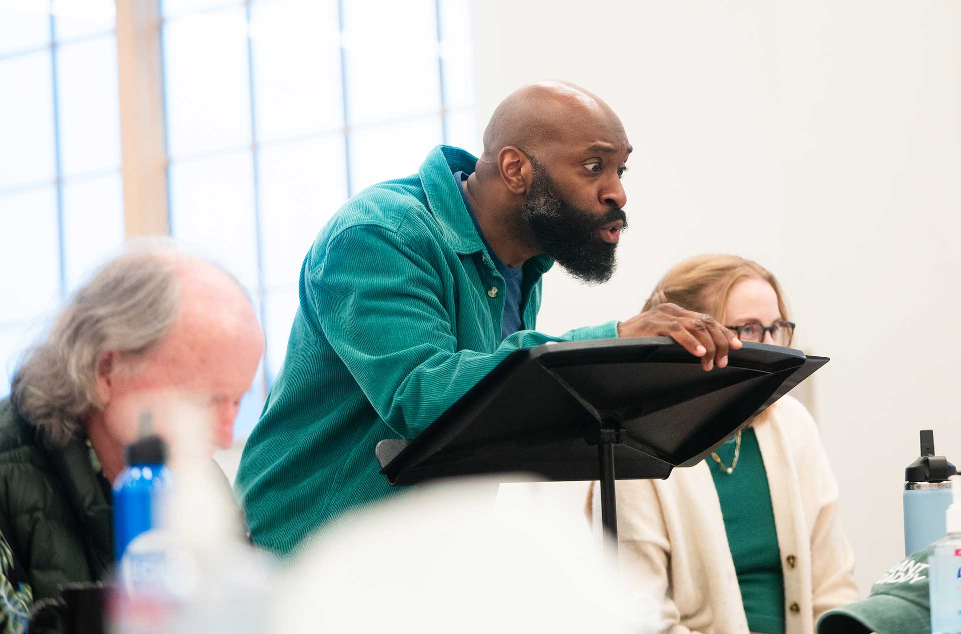 A person in a green corduroy shirt leans over a music stand mid-performance, with an expressive face, while others sit nearby watching.