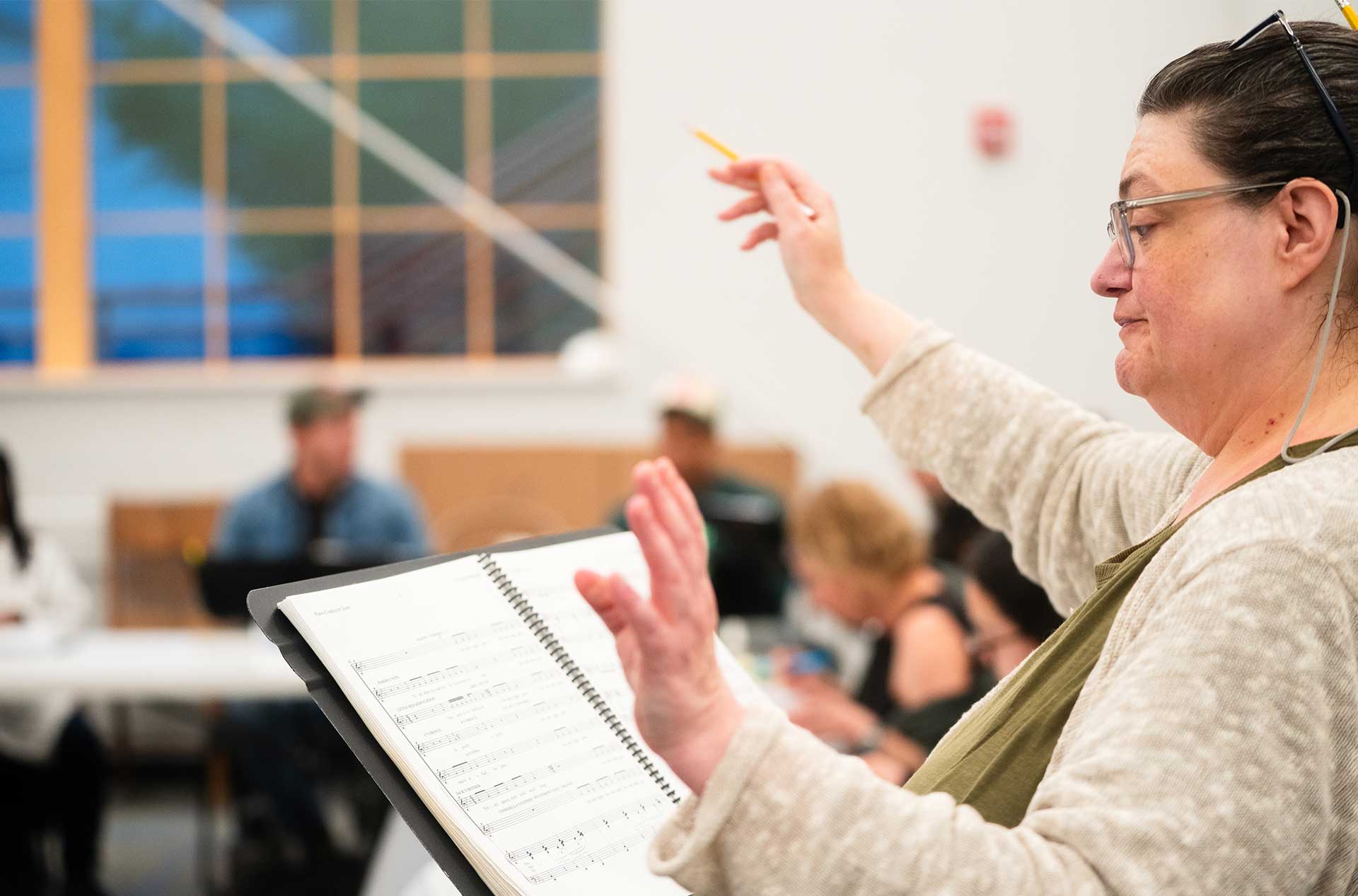 A person gestures mid-conducting with a pencil in one hand and a musical score in the other, focused during rehearsal.