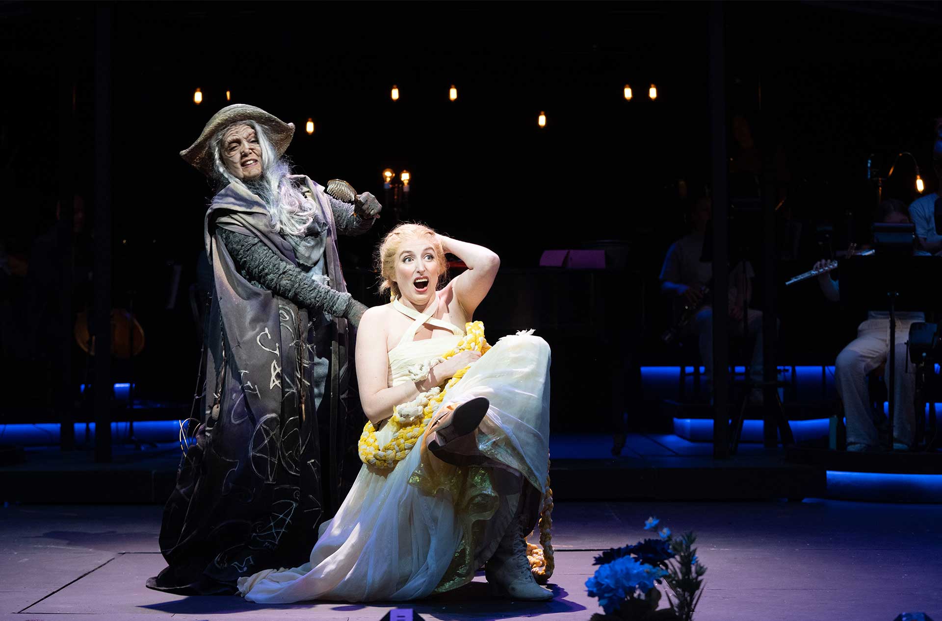 A character in a ragged wizard-like cloak looms over another seated performer in a white and yellow gown with long braided hair, who reacts with a surprised, wide-eyed expression.