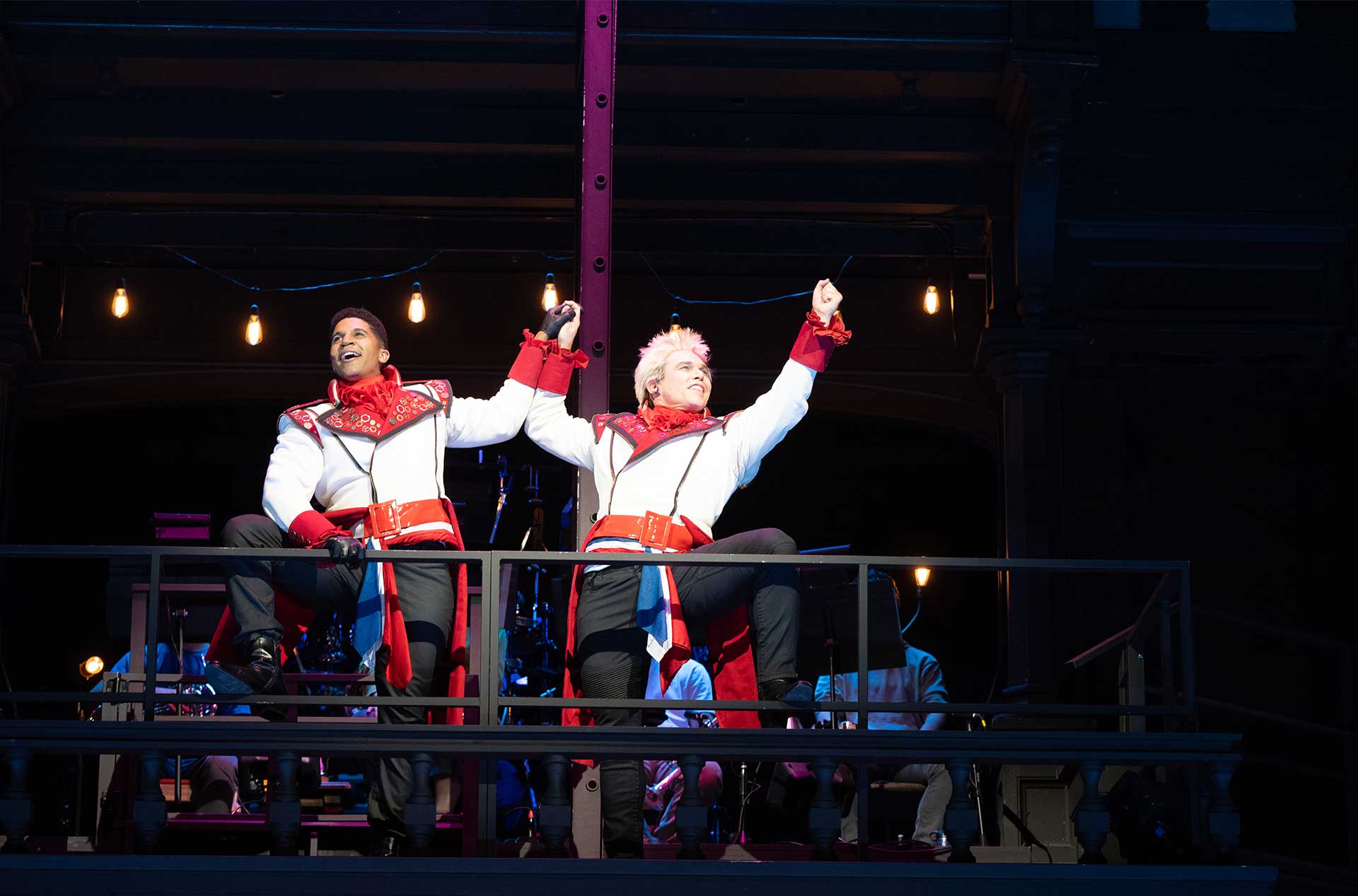 Two actors in identical, flamboyant white and red prince-like costumes strike a triumphant pose on a balcony, holding hands and gesturing boldly.