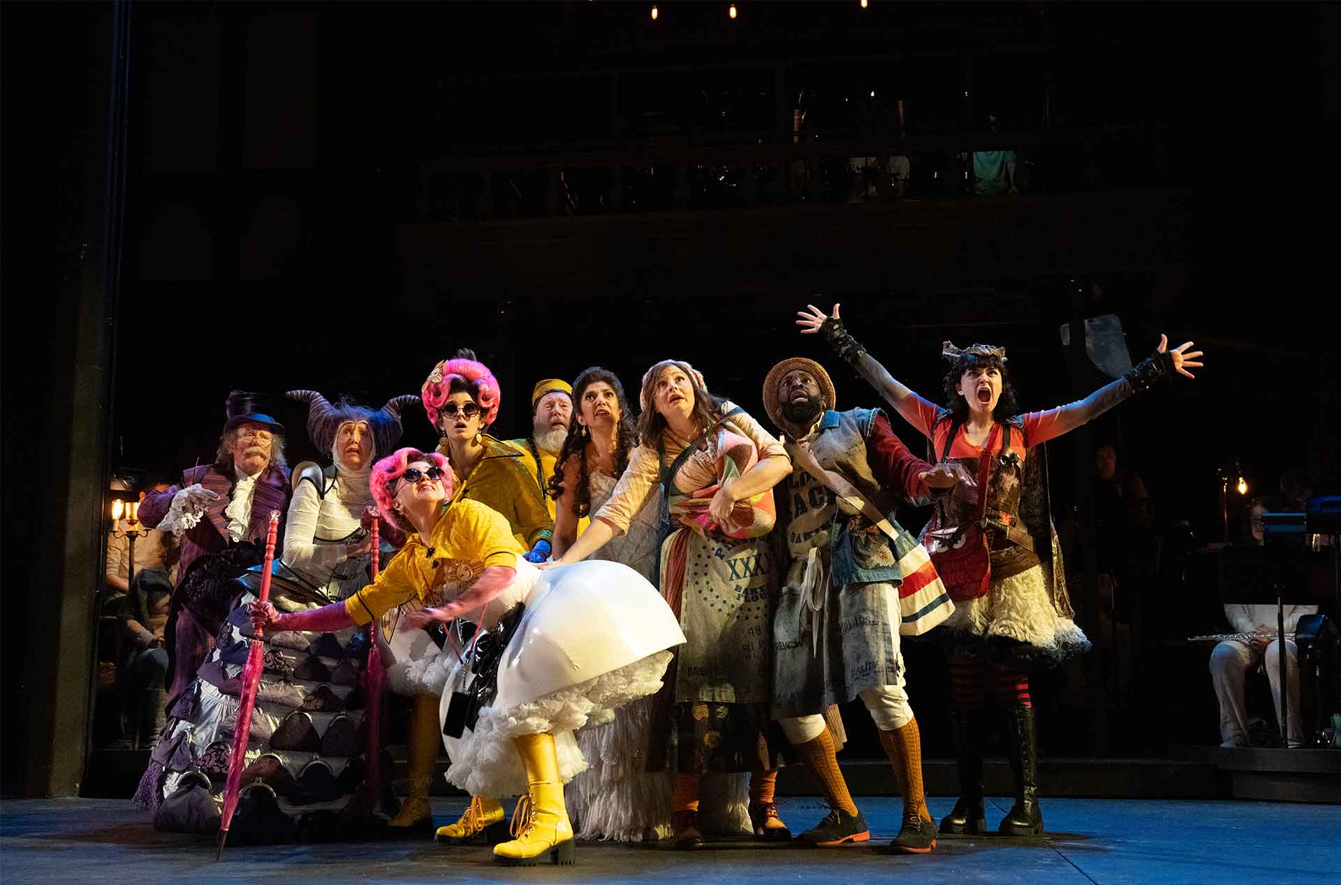 A diverse group of actors in elaborate, whimsical costumes stand tightly huddled on stage, reacting with exaggerated expressions of awe or fear, under warm spotlighting against a dark theater backdrop.