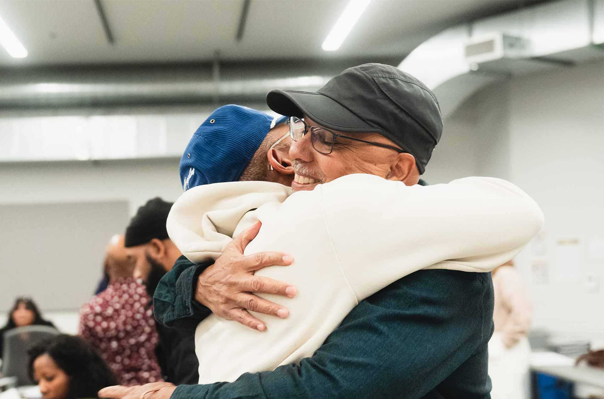 Two people embracing in a warm hug in a bright room, with others in the background.