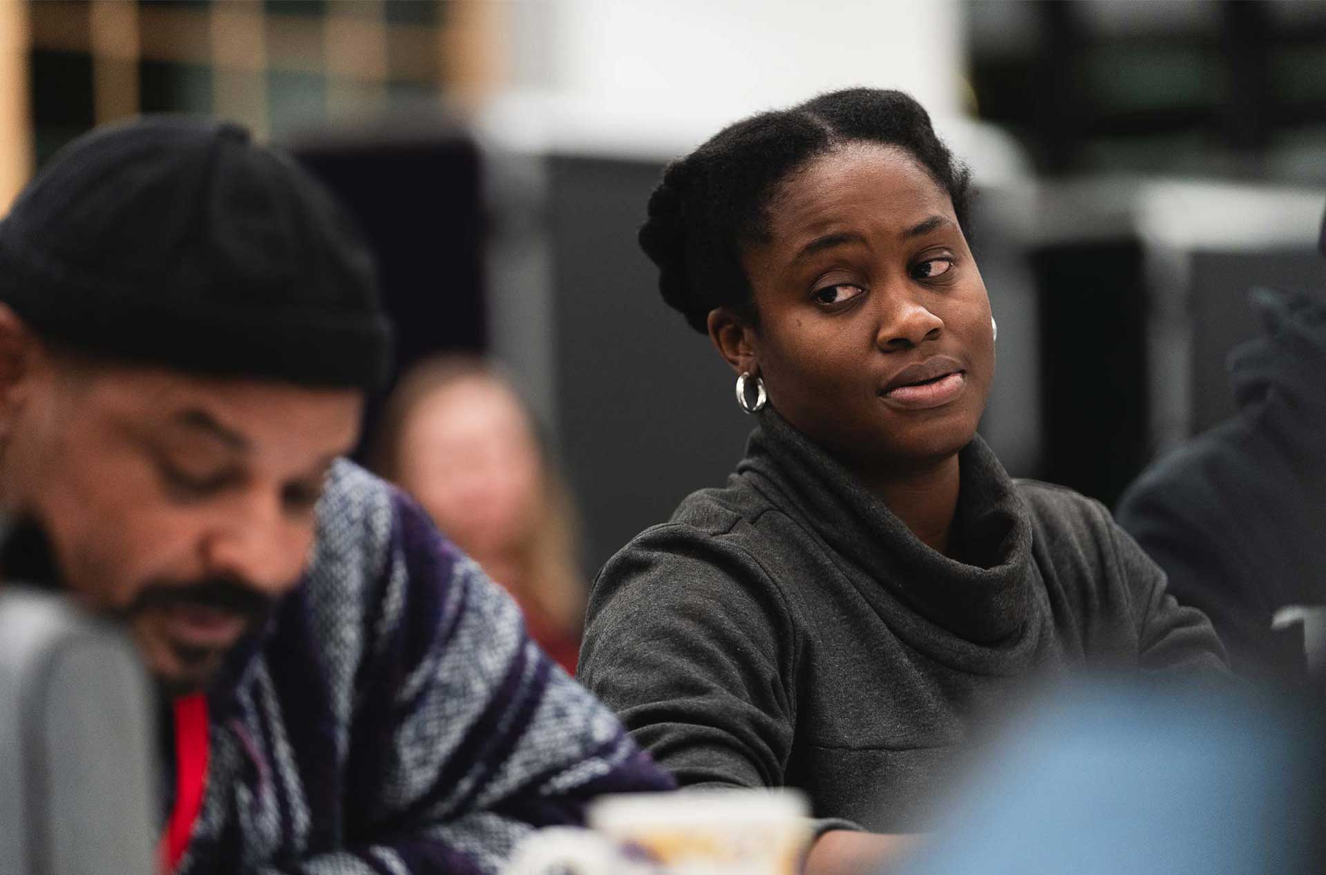 A person wearing a dark sweater listens attentively, glancing sideways during a discussion. Another individual in a patterned sweater sits nearby, focused on writing or reading.