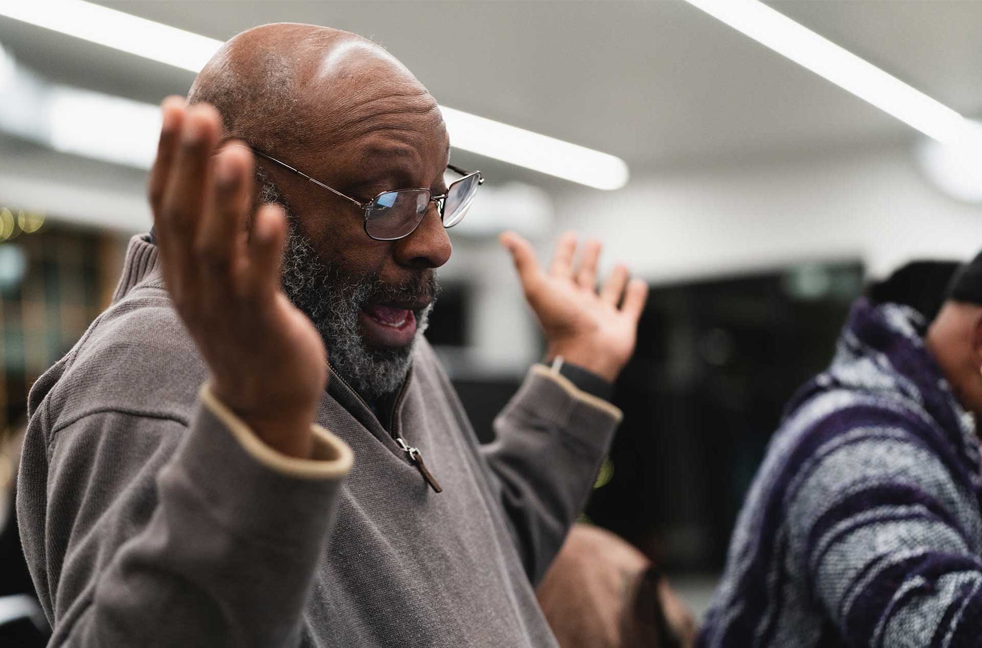A person with glasses gestures with their hands while animatedly speaking at the rehearsal table.